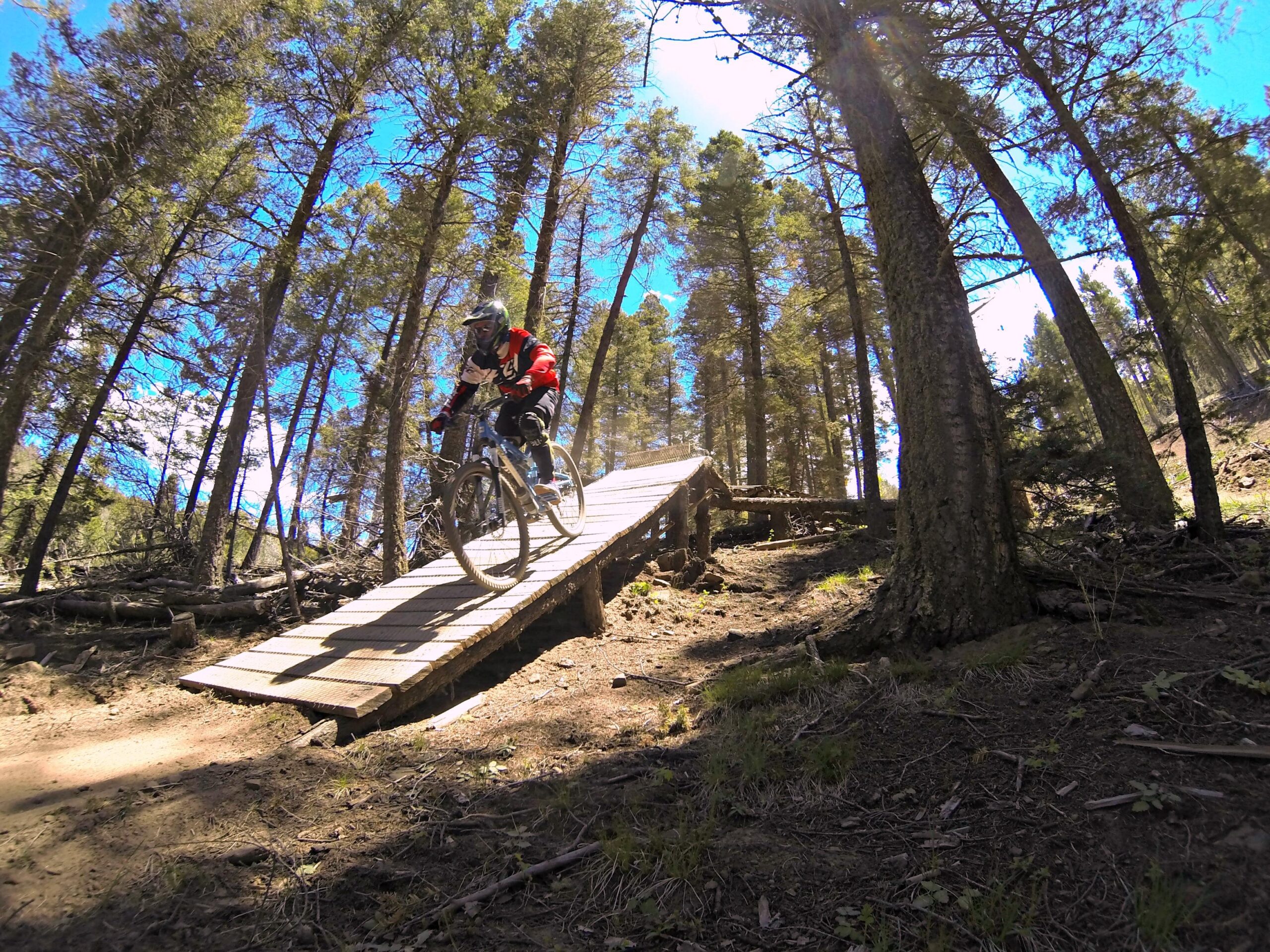A mountain biker rides along a narrow wooden bridge through a forest, surrounded by tall trees under a blue sky. Angel Fire Bike Park mountain bike trail.