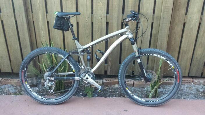 Trek Fuel EX 7: A mountain bike parked on a paved surface in front of a wooden fence. The bike features wide tires and a front suspension, and a small bag is attached to the seat. In the background, there are some plants and a few bricks visible.