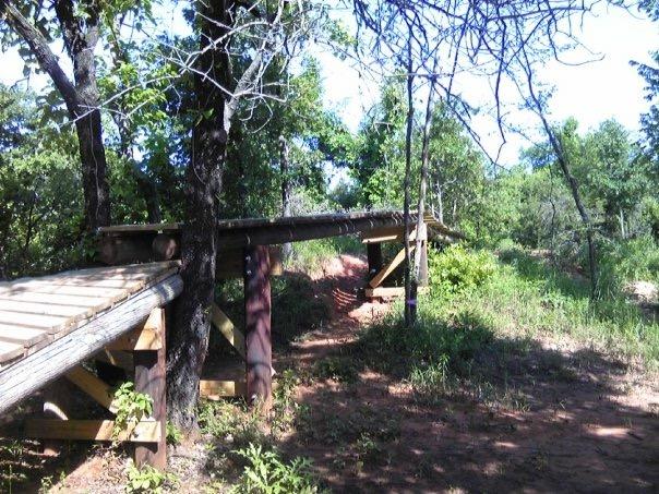 A rustic wooden bridge connecting two paths in a wooded area, surrounded by lush greenery and trees. The bridge is made of logs and has sturdy supports, extending over a natural landscape with soft earth and grass visible along the trail. Sunlight filters through the leaves, creating a serene outdoor atmosphere. Lake Stanley Draper mountain bike trail.