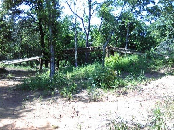 Wooden bridge partially obscured by trees and shrubs in a natural setting. The area is grassy and sandy, with dappled sunlight filtering through the leaves. Lake Stanley Draper mountain bike trail.