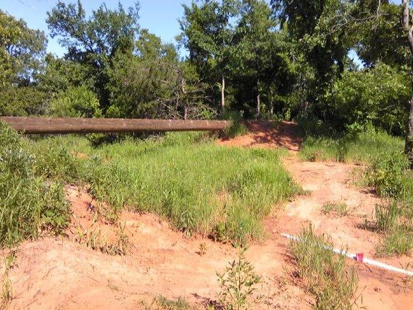 A dirt path surrounded by tall green grass and trees, leading to a wooden beam or post that lies horizontally across the scene. The ground is sandy, indicating a warm, natural setting. Lake Stanley Draper mountain bike trail.