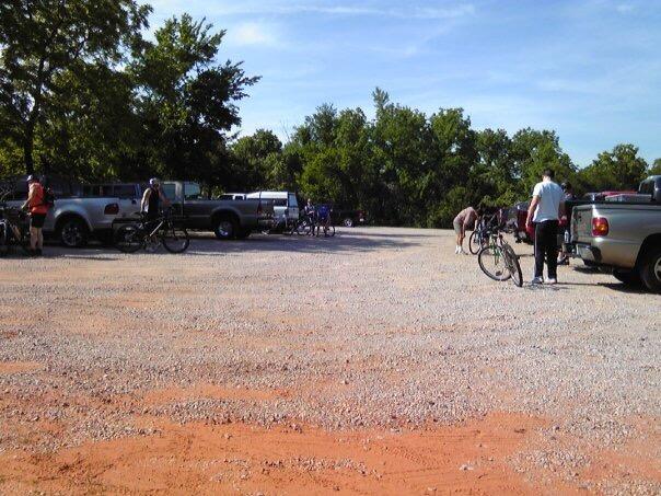 A gravel parking area with several pickup trucks parked on the left, where a group of people is gathered, preparing their bicycles. The scene is set against a backdrop of greenery, with clear skies above. Lake Stanley Draper mountain bike trail.