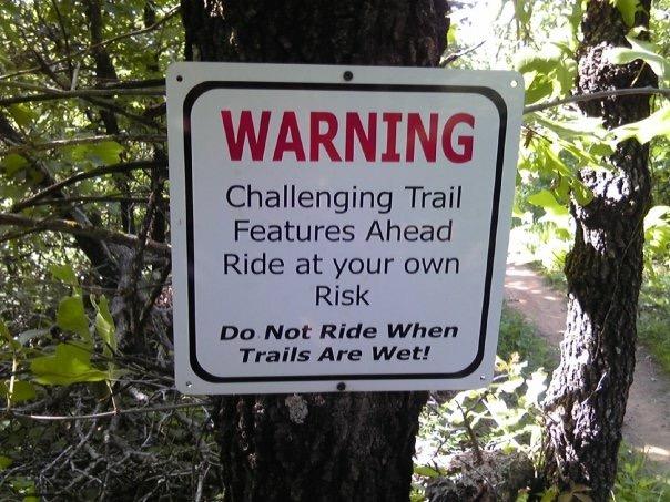 A warning sign attached to a tree in a wooded area, indicating challenging trail features ahead and advising riders to proceed at their own risk. The sign also states, "Do Not Ride When Trails Are Wet!" Lake Stanley Draper mountain bike trail.