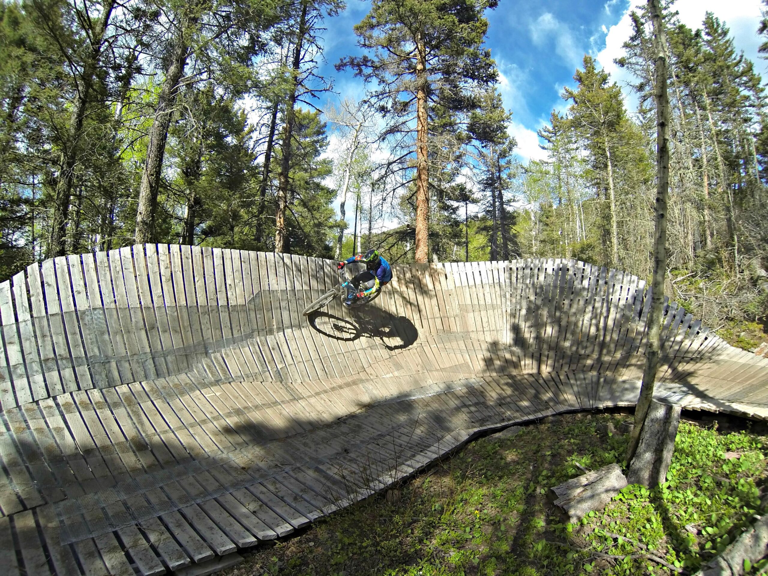 A mountain biker leans into a wooden berm on a forest trail, surrounded by tall trees and bright blue skies, capturing the thrill of downhill riding. Angel Fire Bike Park mountain bike trail.
