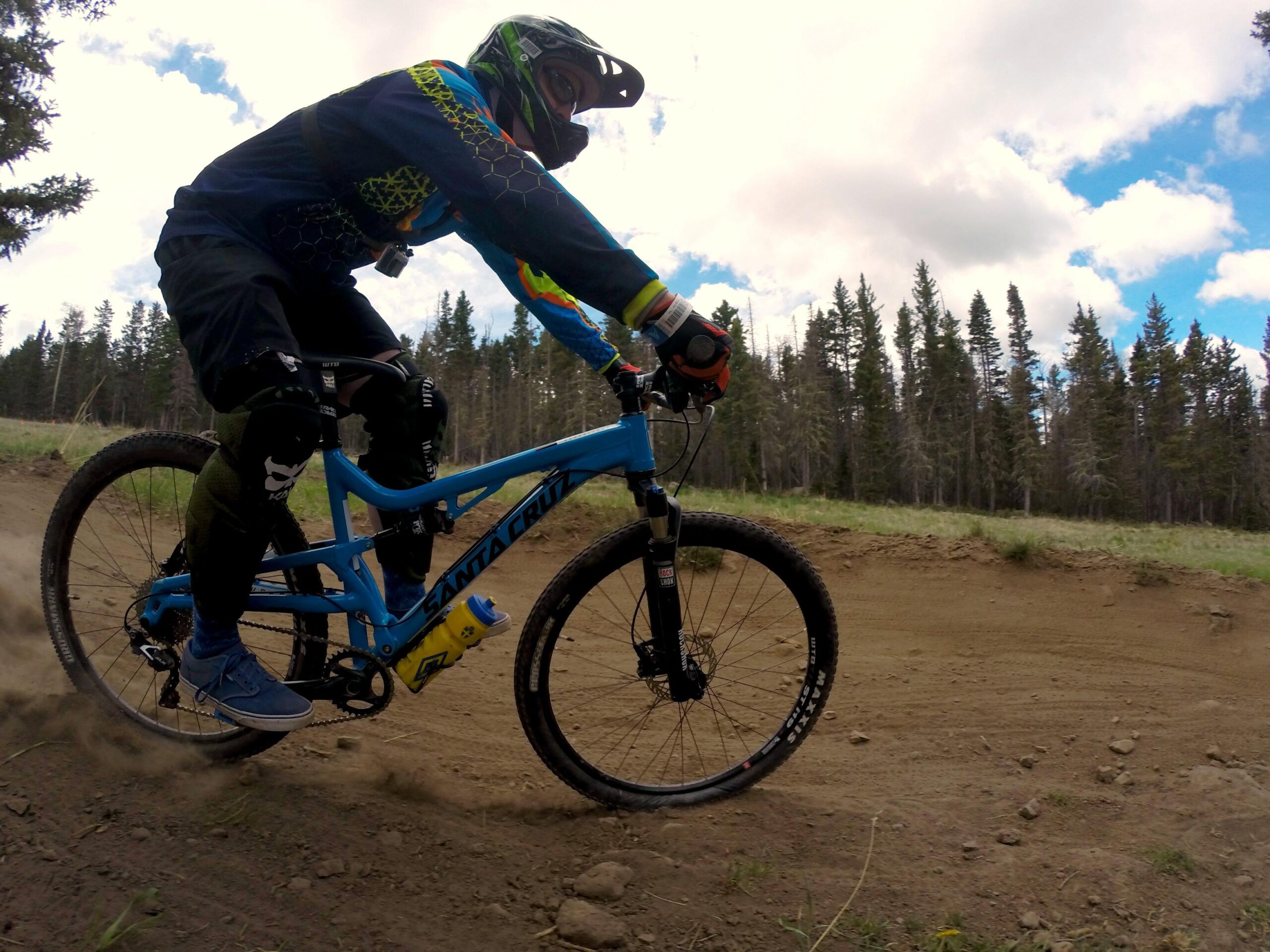Santa Cruz Superlight: A mountain biker in protective gear rides on a dirt trail surrounded by trees. The bike is blue with a prominent brand logo, and the rider is leaning into a turn, leaving a trail of dust behind. The sky is partly cloudy, indicating an outdoor setting.
