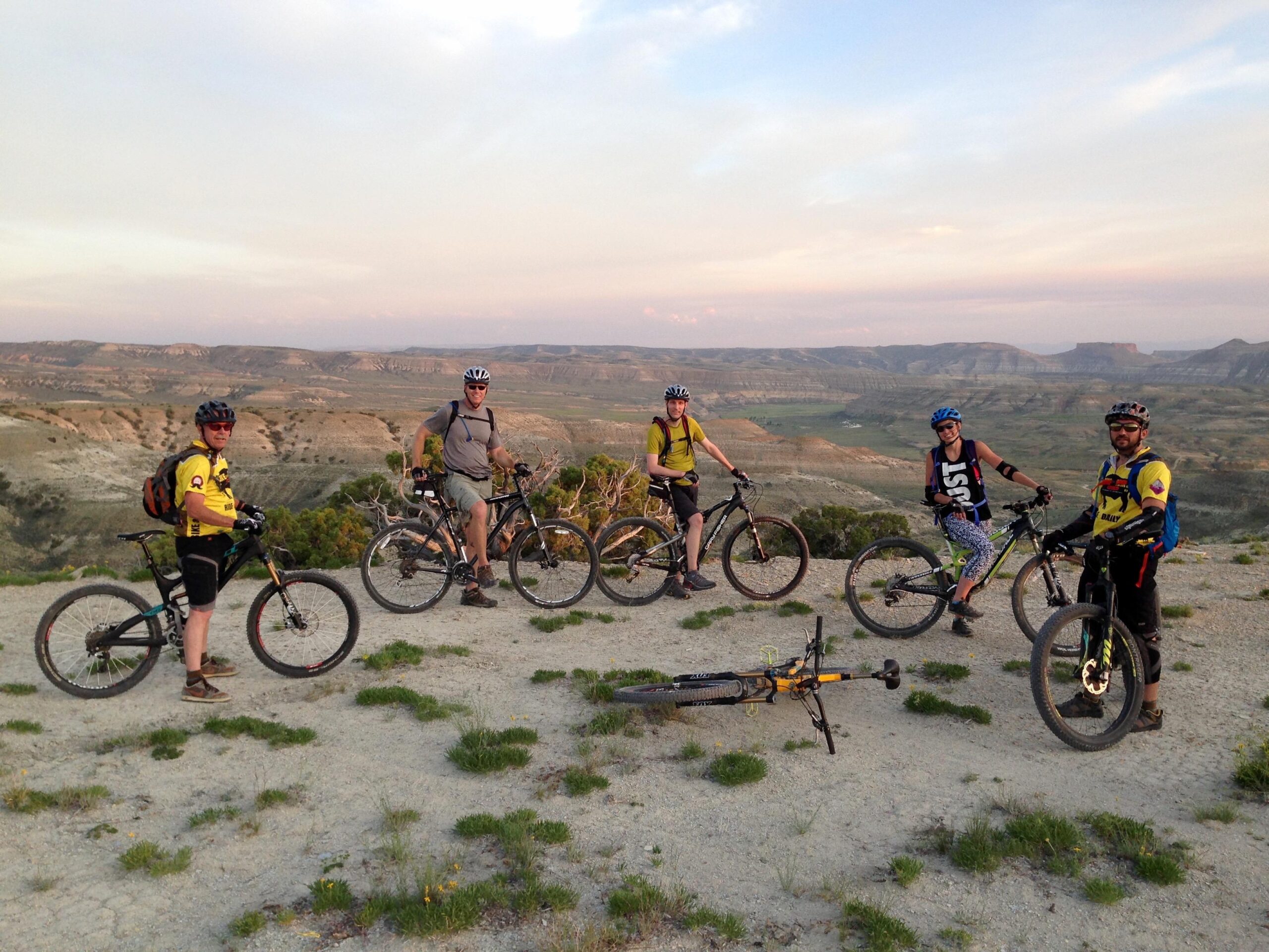 A group of six mountain bikers poses on a rocky overlook with a scenic view of rolling hills and valleys in the background. The cyclists, wearing helmets and brightly colored jerseys, stand beside their bikes, enjoying the sunset. One bike is lying on the ground. The landscape features a mixture of shrubs and barren terrain, highlighting a beautiful outdoor adventure setting. Wilkins Peak Trails mountain bike trail.