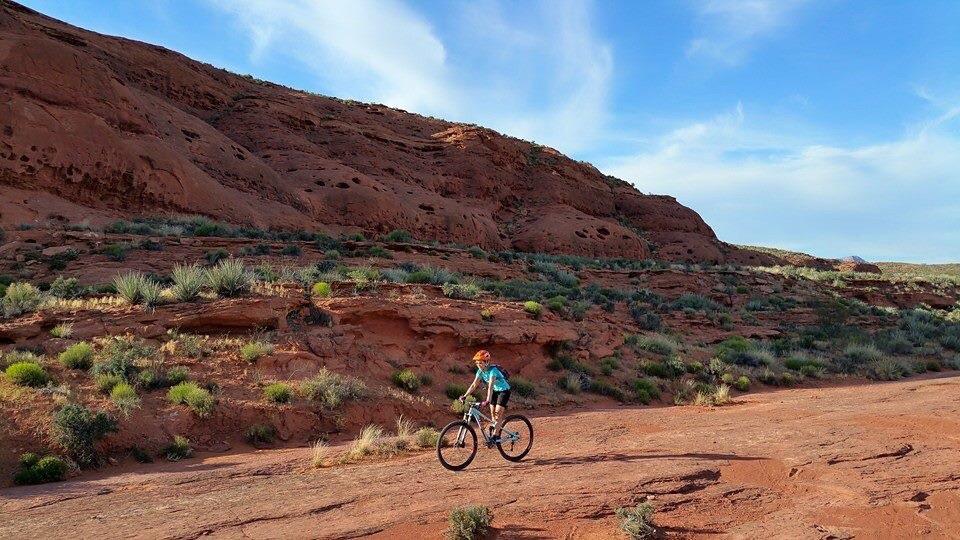 A person riding a bicycle on a rocky trail surrounded by red sandstone cliffs and sparse green vegetation under a blue sky. Prospector - Church Rocks Loop mountain bike trail.