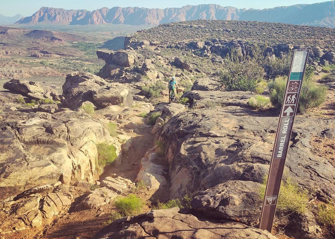 A mountain biker rides along a rocky trail surrounded by desert vegetation, with distant mountains in the background. A trail sign labeled "Barrel Roll" indicates the path ahead. The landscape features textured rocks and sparse greenery under a clear sky. Barrel Roll mountain bike trail.