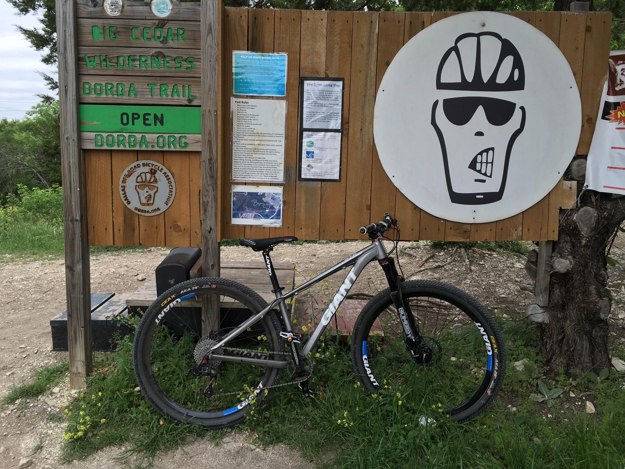 Giant XTC 29er 2: A mountain bike leaning against a wooden information sign at the Big Cedar Wilderness Dorba Trail. The sign displays text indicating the trail is open and features various park rules and informational posters. A distinctive circular logo with a skull wearing sunglasses and a bike helmet is visible on the sign. Surrounding vegetation includes grass and small yellow flowers.
