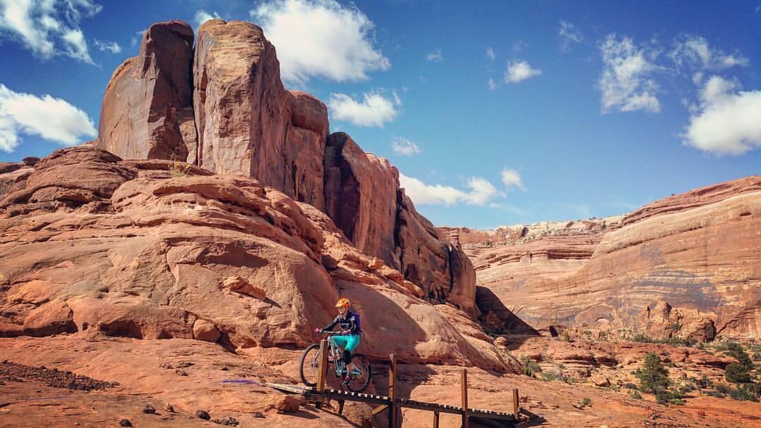 A mountain biker navigating a wooden bridge on a rocky terrain, with towering red rock formations and a blue sky dotted with clouds in the background. Navajo Rocks mountain bike trail.