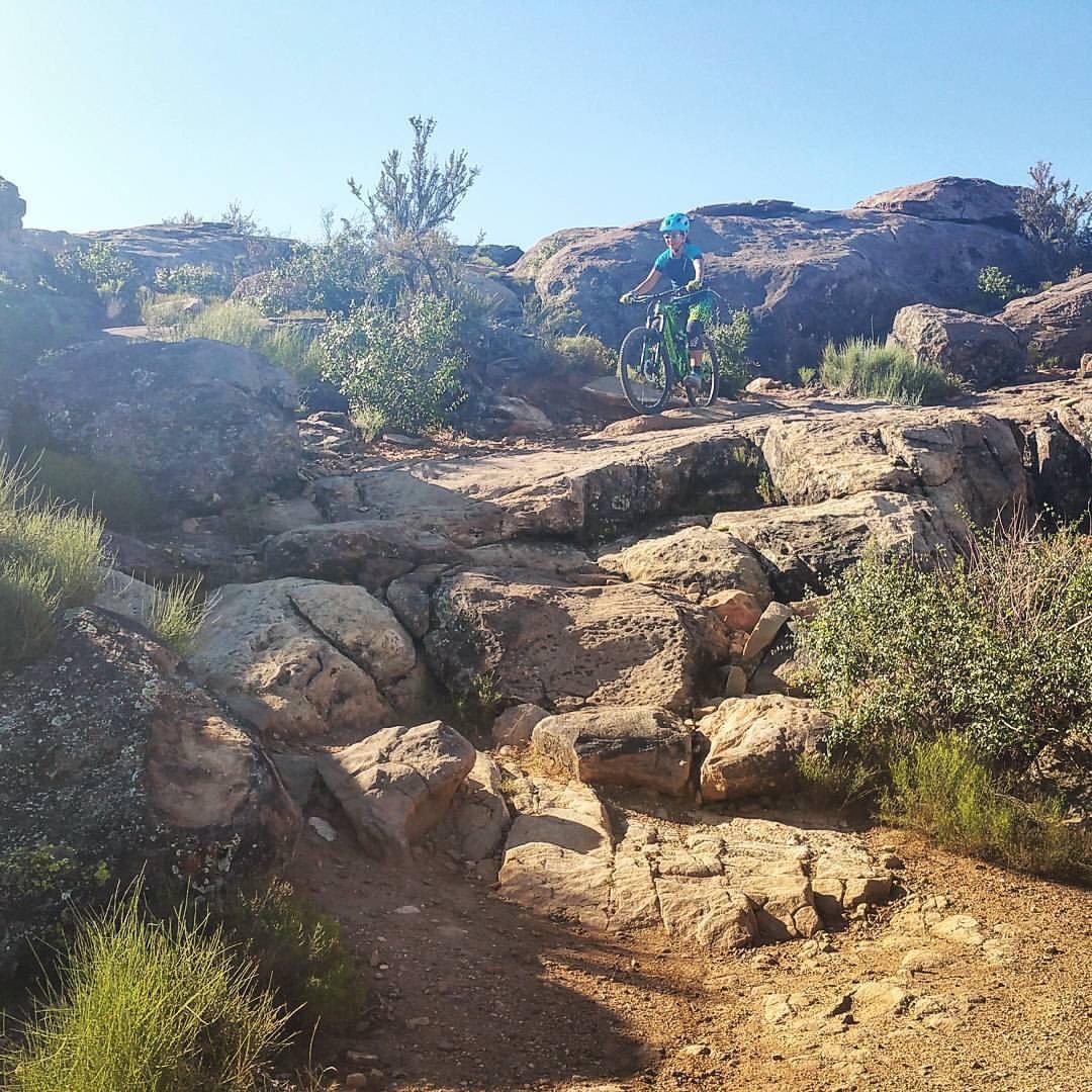 A person riding a mountain bike on a rocky trail surrounded by shrubs and boulders under a clear blue sky. Barrel Roll mountain bike trail.