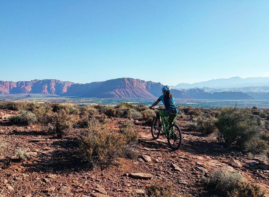A person riding a mountain bike on a rocky trail, with a backdrop of red rock mountains and a clear blue sky. The rider is wearing a helmet and colorful cycling attire, and the landscape features sparse vegetation typical of desert terrain. Barrel Roll mountain bike trail.
