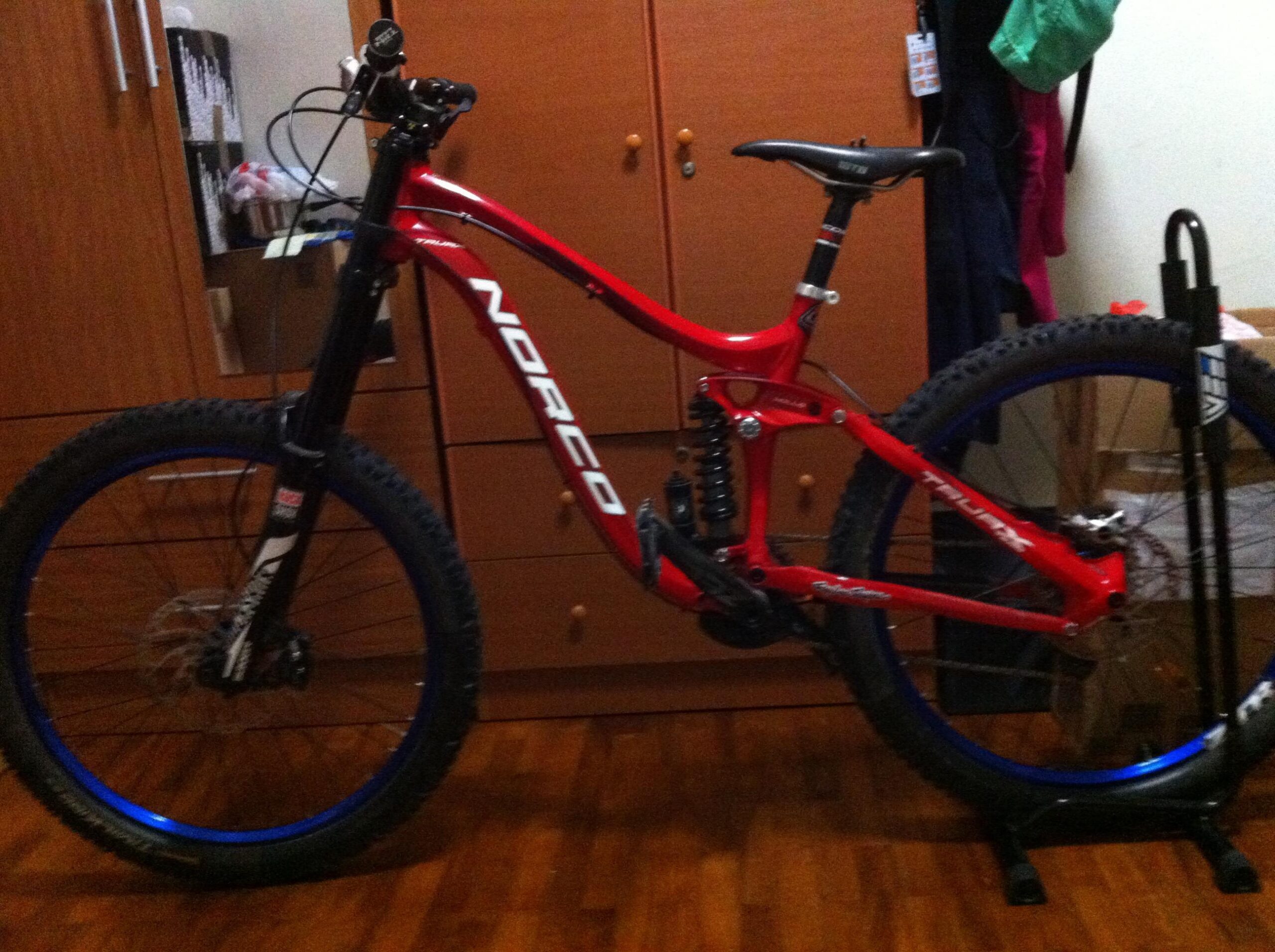 Norco TRUAX 1: A red Norco mountain bike with blue rims, standing on a bike stand against a backdrop of wooden furniture and a cluttered room.