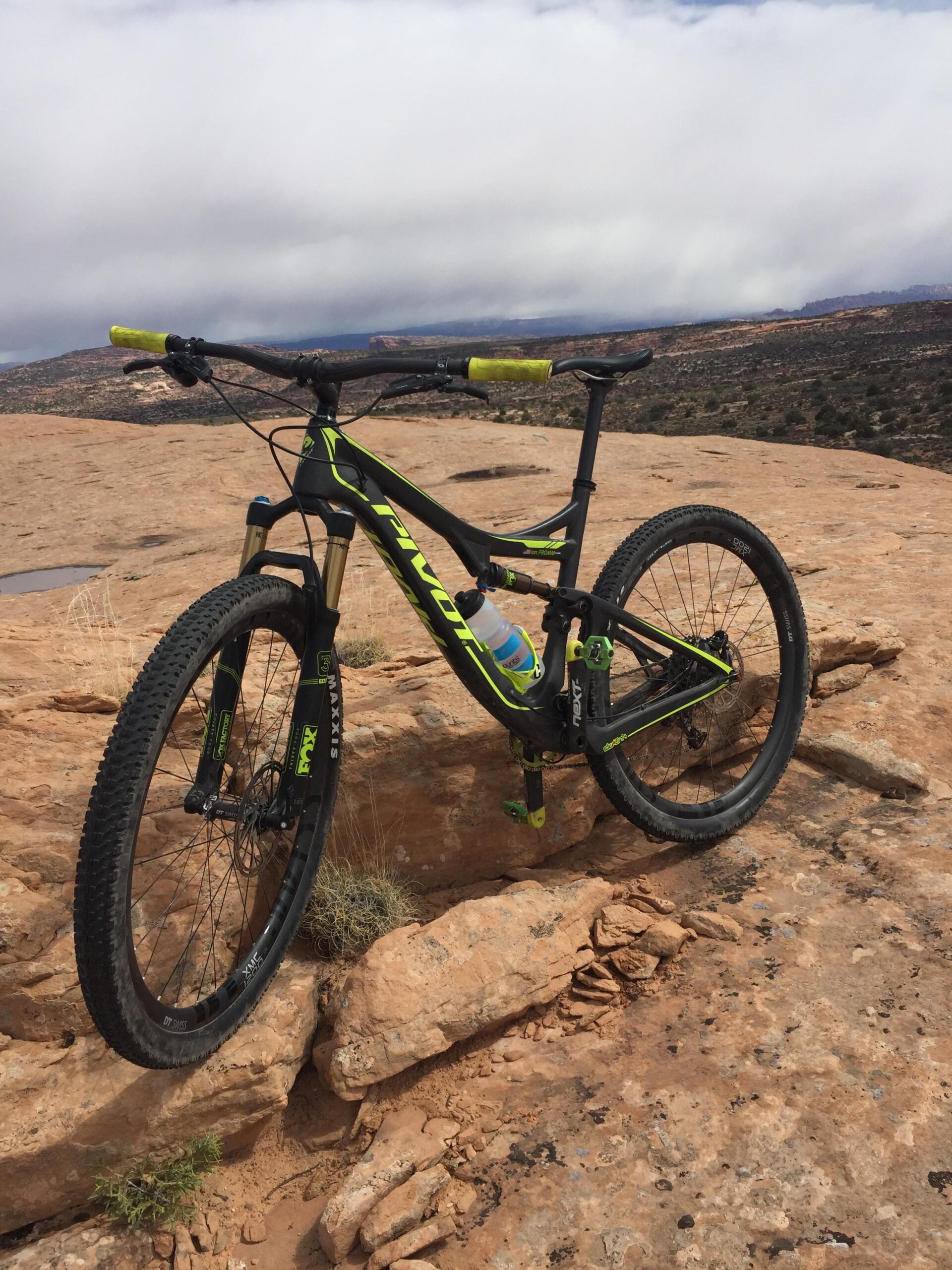 Pivot Mach 429SL Carbon: A mountain bike resting on rocky terrain, with a scenic backdrop featuring rolling hills and cloudy skies. The bike has a sleek black and lime green design, showcasing its handlebars, front suspension, and wheels.