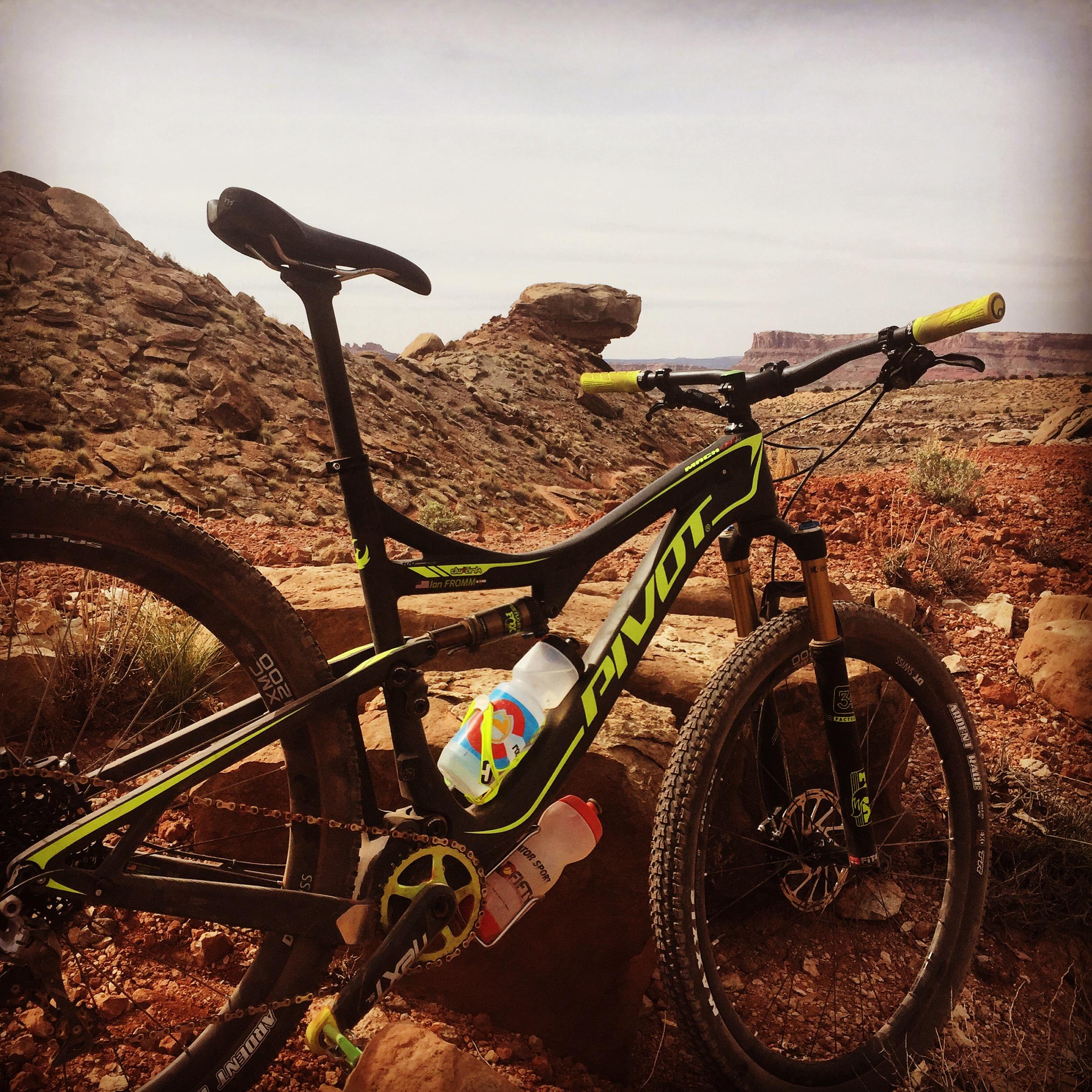 Pivot Mach 429SL Carbon: A mountain bike leaning against a rocky outcrop in a desert landscape, featuring a black and neon green frame with yellow grips. A water bottle is mounted on the bike, and the background includes distant rock formations and a clear sky.