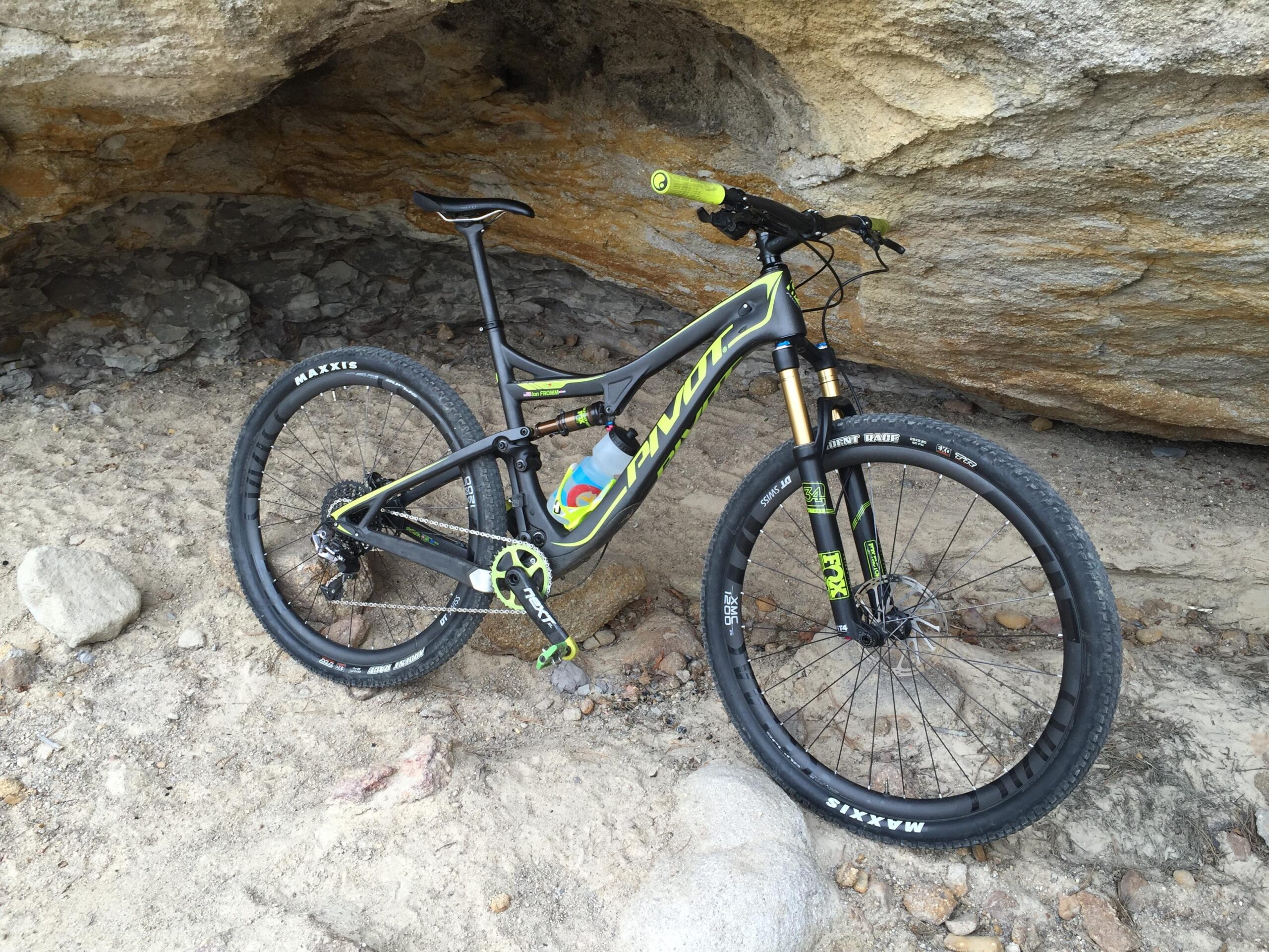 Pivot Mach 429SL Carbon: A mountain bike with a black frame and bright green accents is positioned on sandy ground near a rock overhang. The bike features wide tires, a water bottle holder, and front suspension, highlighting its suitability for off-road terrain.