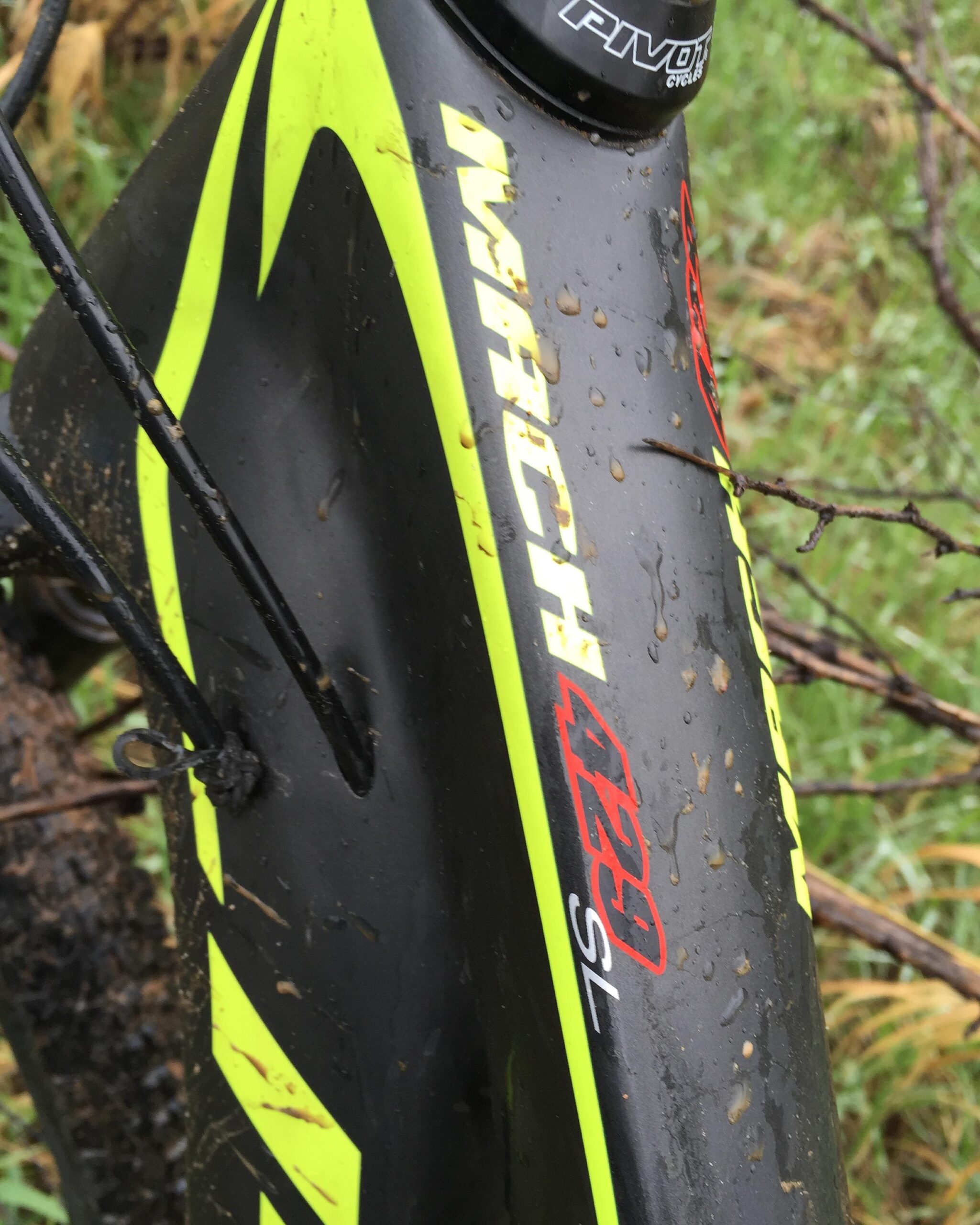 Pivot Mach 429SL Carbon: Close-up of a mountain bike frame showing a black and neon yellow design, with the text "IMPACT 429 SL" visible. The frame is wet, indicating recent exposure to rain or mud, with visible droplets and dirt along the surface. Background features blurred greenery and twigs, suggesting an outdoor setting.