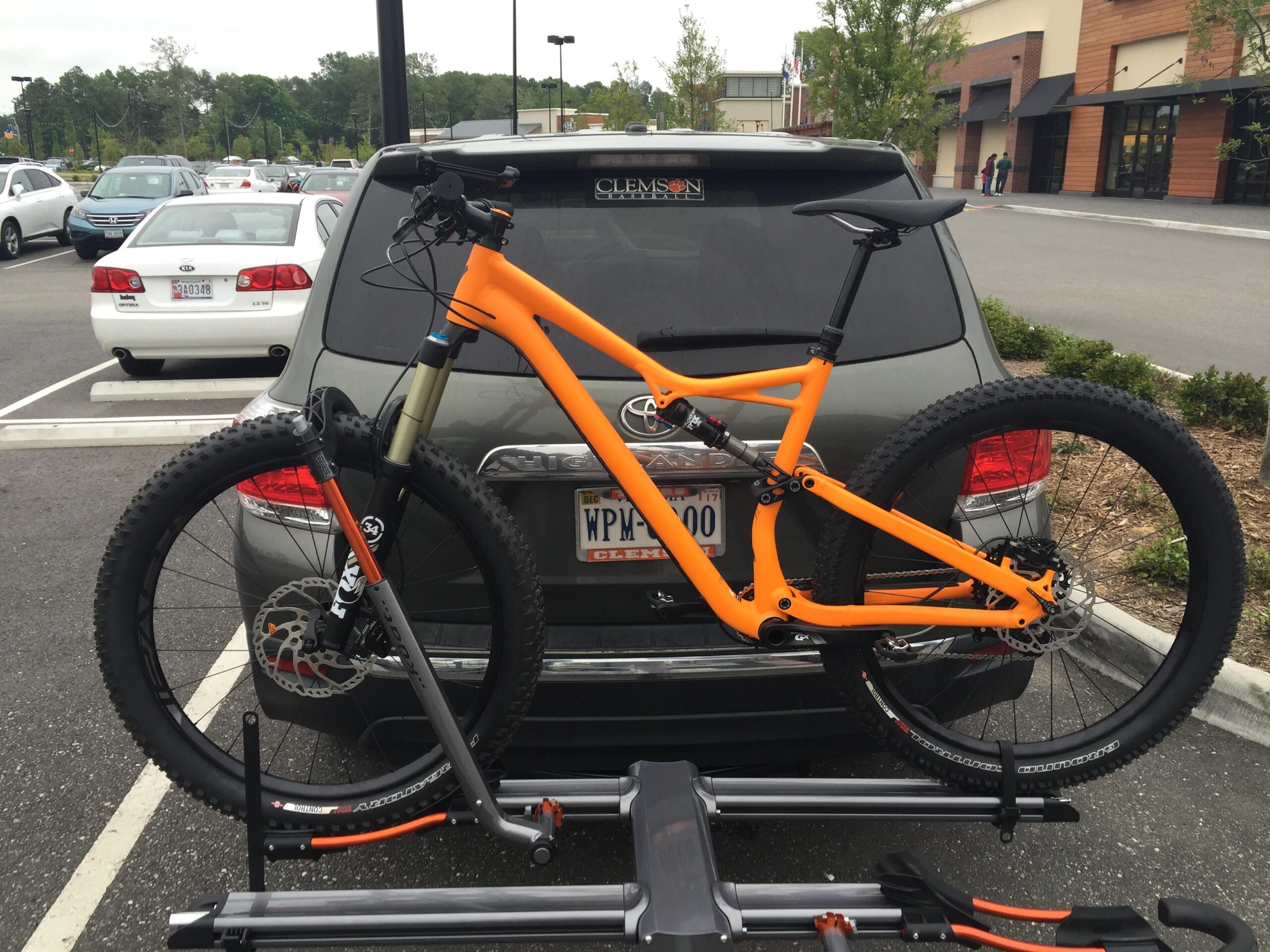 Specialized Stumpjumper FSR 6Fattie: A vibrant orange mountain bike is securely mounted on a bike rack attached to the back of a gray SUV parked in a lot. The scene captures several parked cars in the background and a retail building nearby.