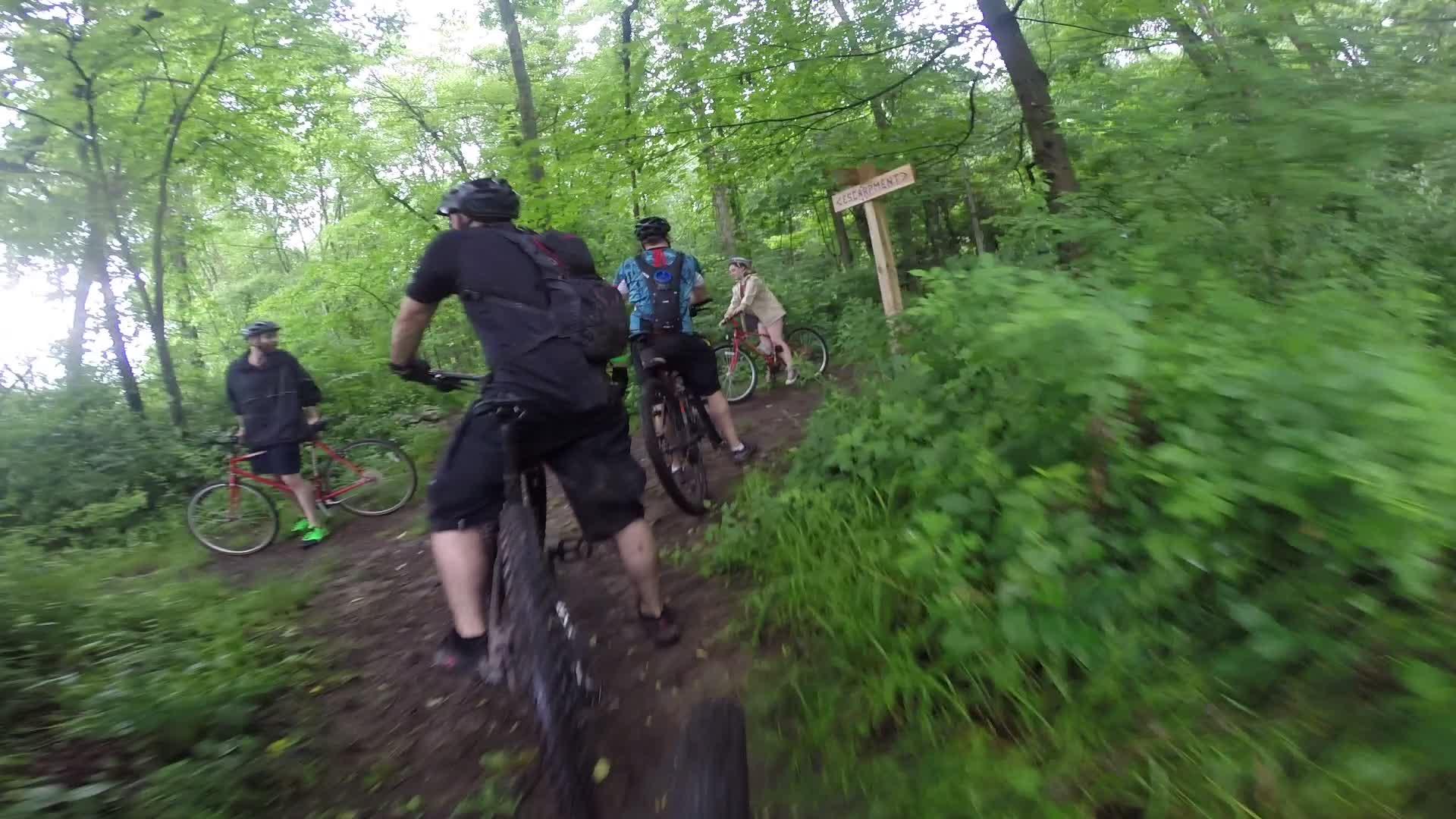A group of cyclists navigating a muddy, wooded trail surrounded by lush green foliage. One cyclist is stopped, facing the camera, while others are in the background, some riding their bikes. A wooden sign visible in the background indicates a nearby destination, adding to the outdoor adventure atmosphere. Stewart State Forest mountain bike trail.