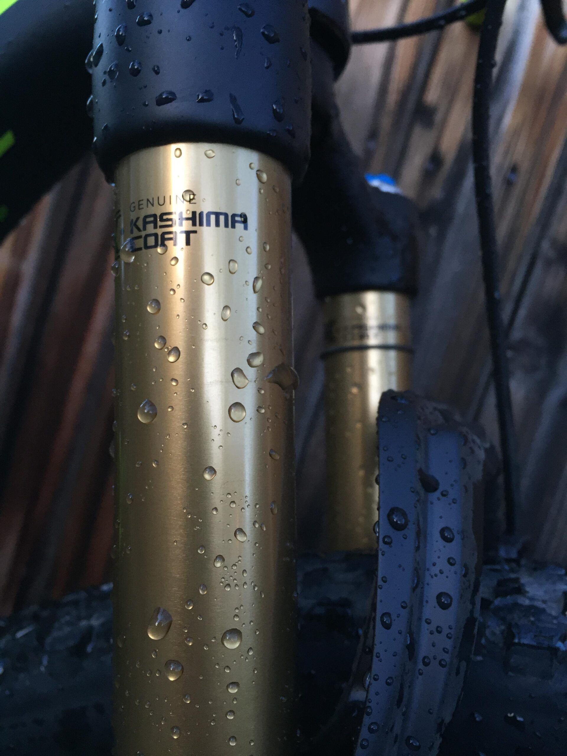 Pivot Mach 429SL Carbon: Close-up of a mountain bike's front suspension fork with "Genuine Kashima Coat" branding. The gold color is accentuated by water droplets, indicating recent exposure to moisture. The fork is partially obscured by the bike's black tire and visible details highlight the textured grip of the handlebar. Background features soft wooden textures.