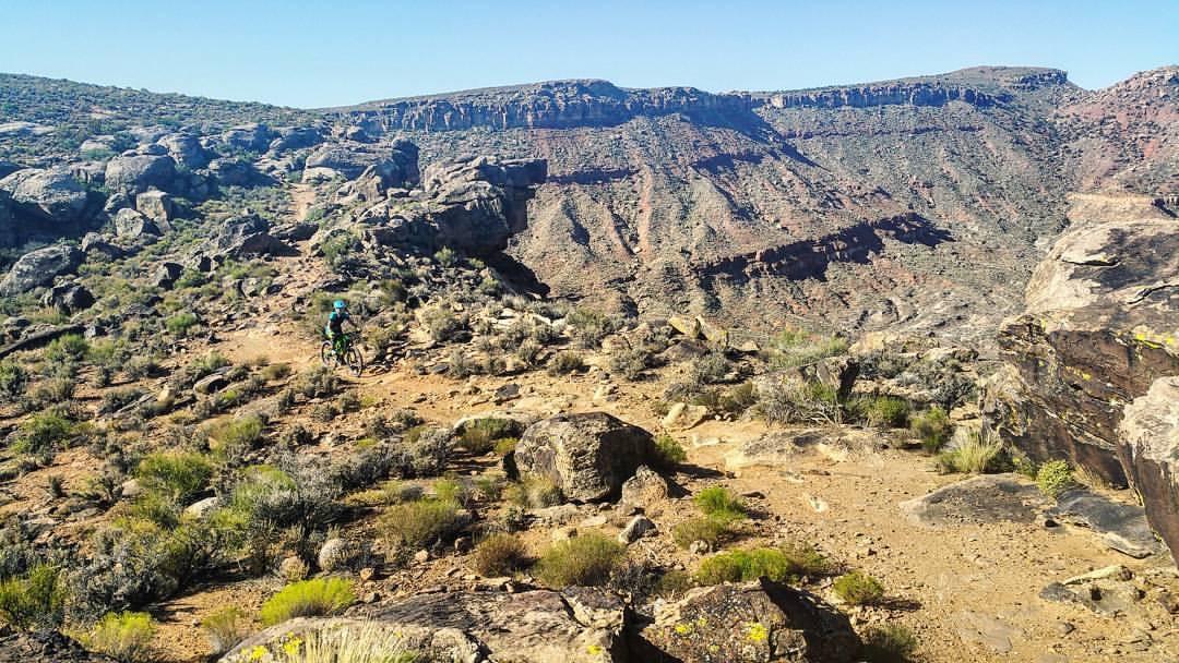 A mountain biker navigates a rugged dirt trail surrounded by rocky terrain and sparse vegetation, with a steep canyon in the background under a clear blue sky. Barrel Roll mountain bike trail.