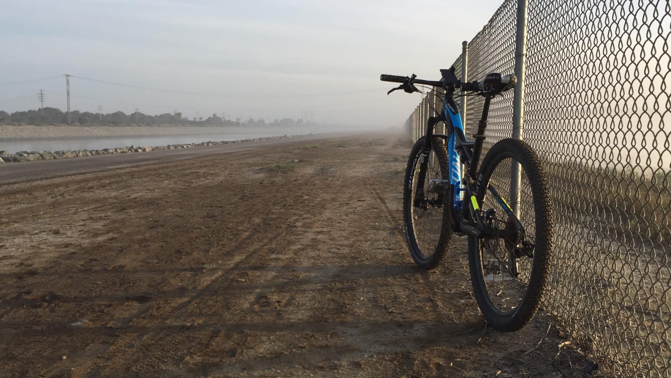 Specialized Stumpjumper FSR: A mountain bike leaning against a chain-link fence along a dirt path, with a misty river and power lines in the background. The scene is set in early morning light, highlighting the quiet and serene atmosphere of the outdoor surroundings.