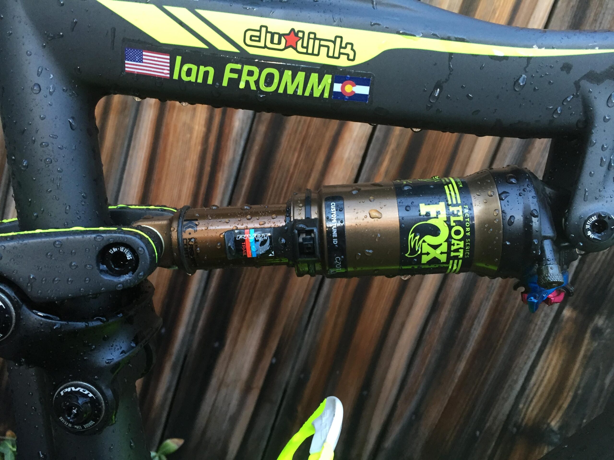 Pivot Mach 429SL Carbon: Close-up image of a mountain bike shock absorber, featuring raindrops on the surface. The shock is branded with "FOX Float" and includes a custom color scheme. The bike frame displays the name "Ian FROMM," along with U.S. and Colorado flags. The background consists of wooden texture.