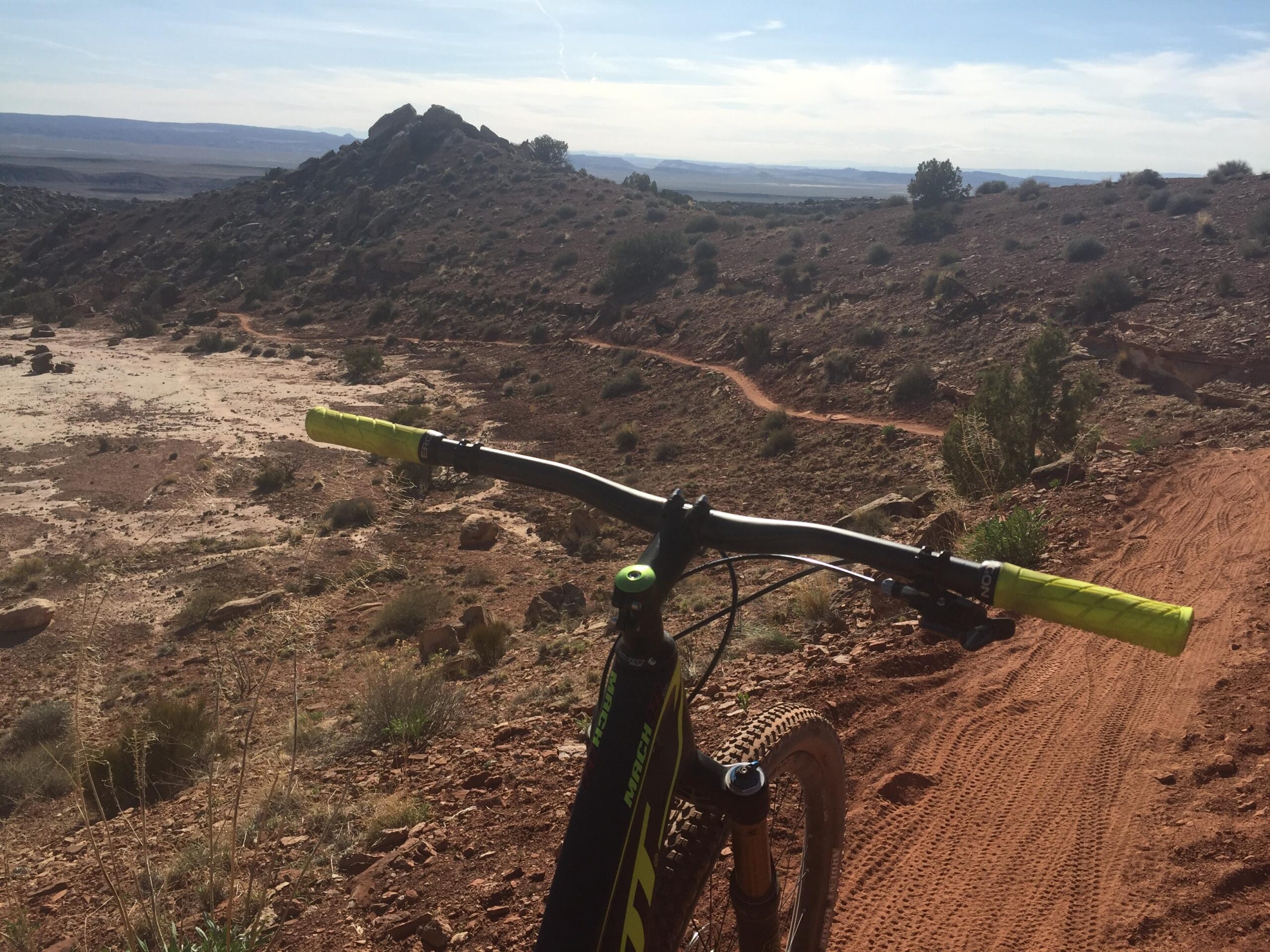 Pivot Mach 429SL Carbon: A view from the handlebars of a mountain bike, overlooking a rocky trail that winds through a desert landscape. The terrain features reddish soil and sparse vegetation, with distant hills and blue sky in the background.