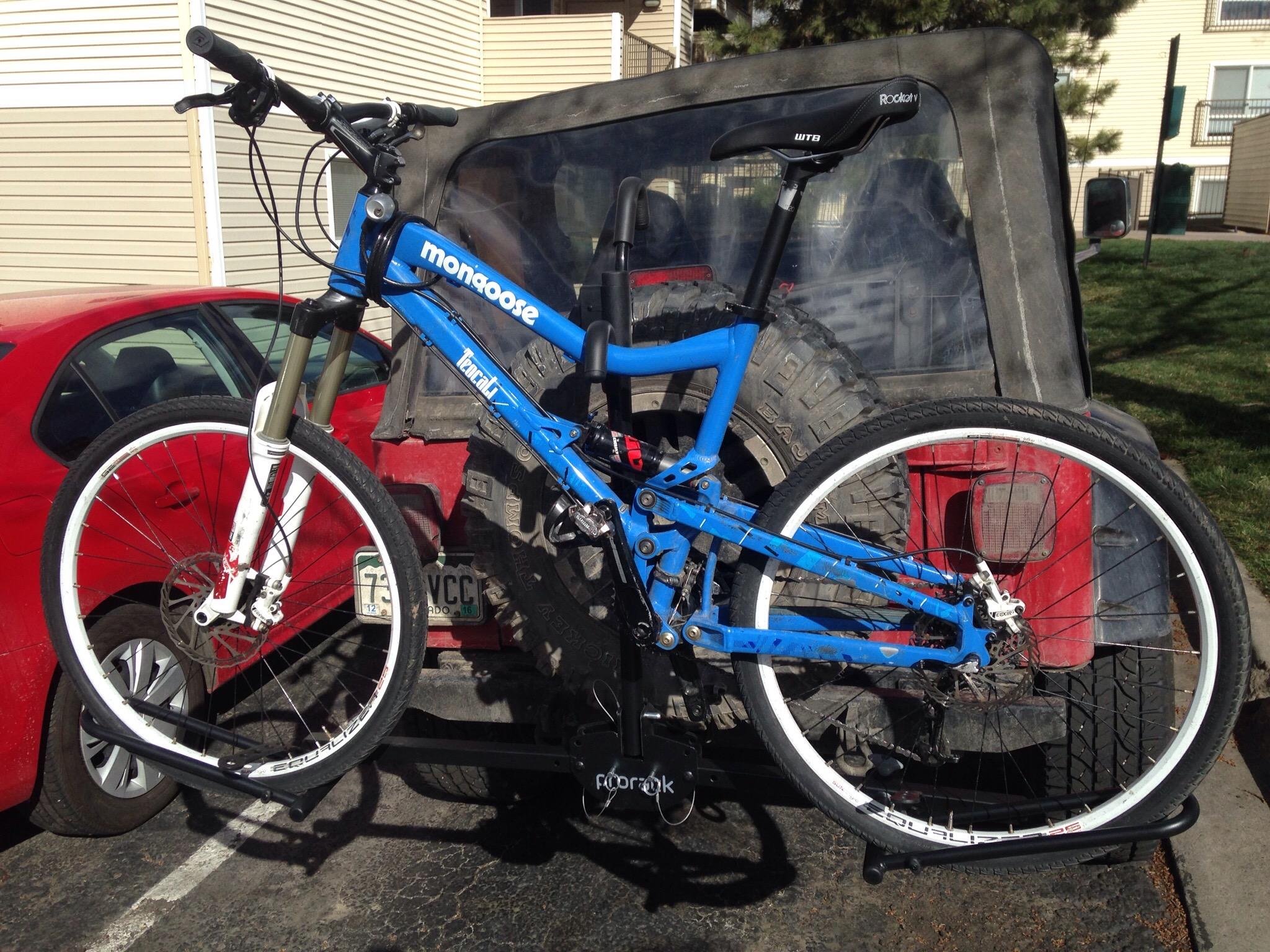 Mongoose Teocali Comp: A blue mountain bike mounted on a bike rack attached to the back of a red car, parked in a lot with residential buildings in the background. The bike features front suspension and white wheels, while a Jeep with a covered trunk is also visible behind the car.