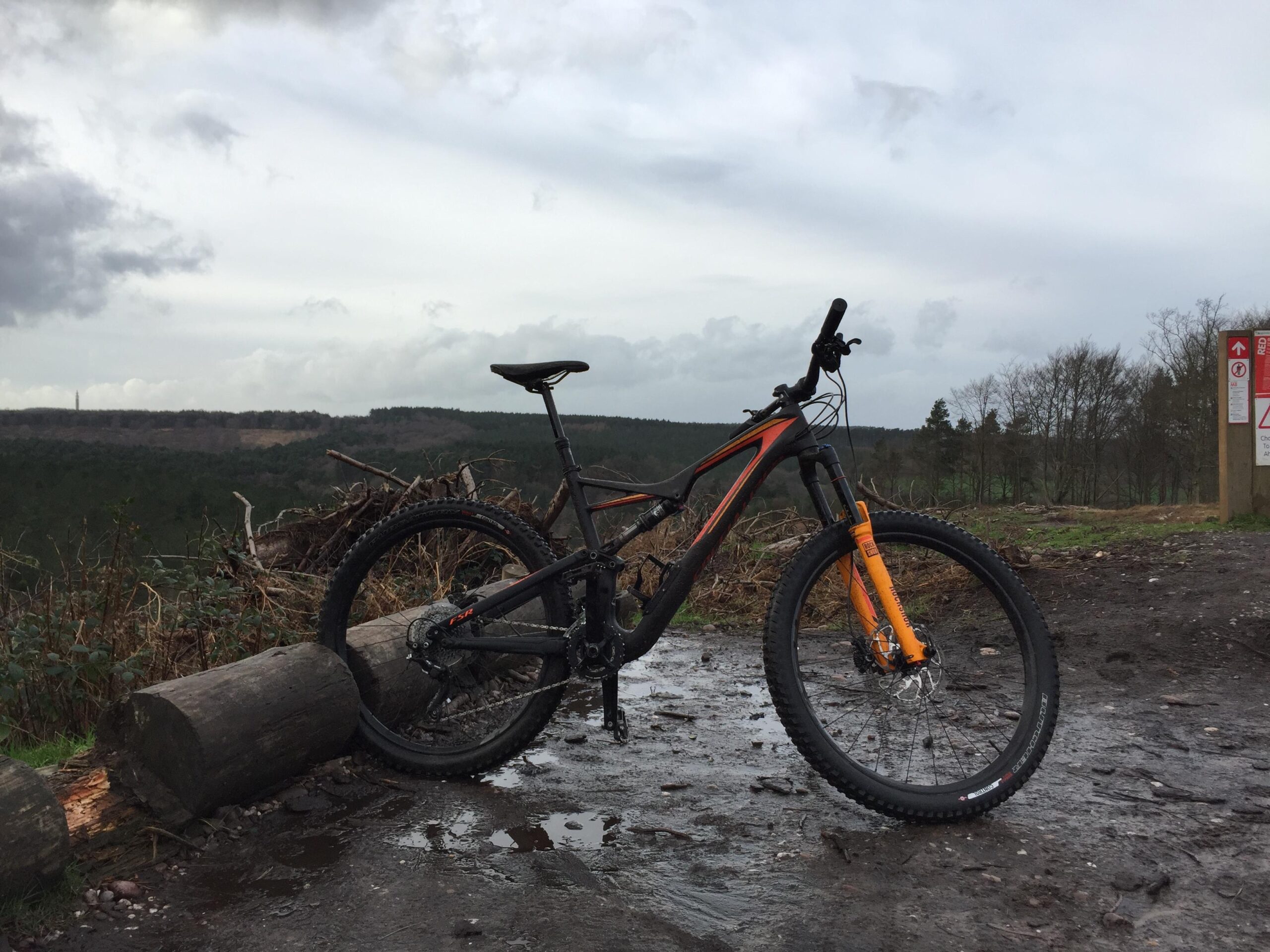 Specialized Stumpjumper FSR: A black and orange mountain bike resting against a log on a muddy trail, with a forested hillside in the background and overcast skies above. A sign is visible in the right side of the image, indicating trail information.