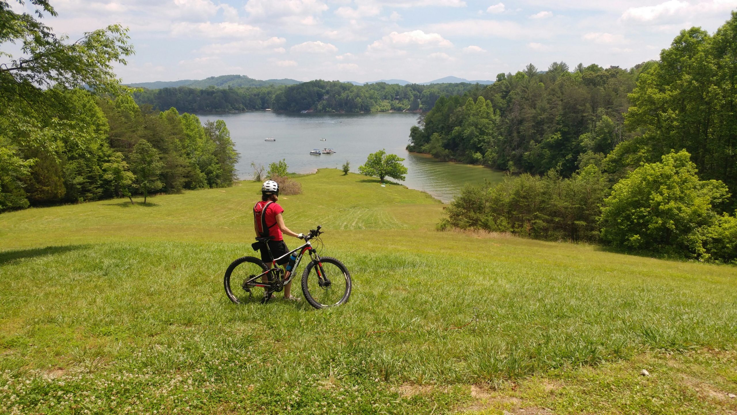 A person wearing a helmet and a red shirt stands with a mountain bike on a grassy hill overlooking a calm lake surrounded by trees. In the background, several boats can be seen on the water, with rolling hills and a partly cloudy sky in the distance. Warrior Creek mountain bike trail.