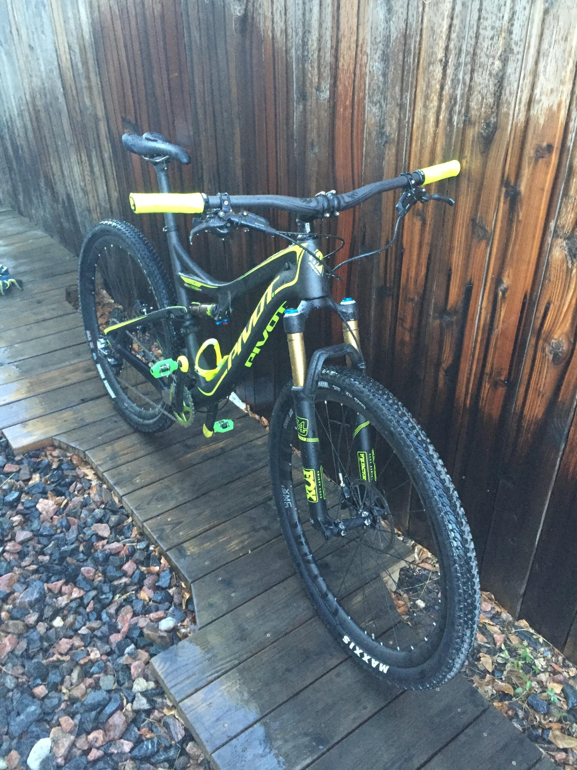 Pivot Mach 429SL Carbon: A black and yellow mountain bike leaning against a wooden fence, positioned on a wooden pathway surrounded by rocks. The bike features knobby tires and vibrant green accessories, with the frame prominently displaying the brand name "Pivot."