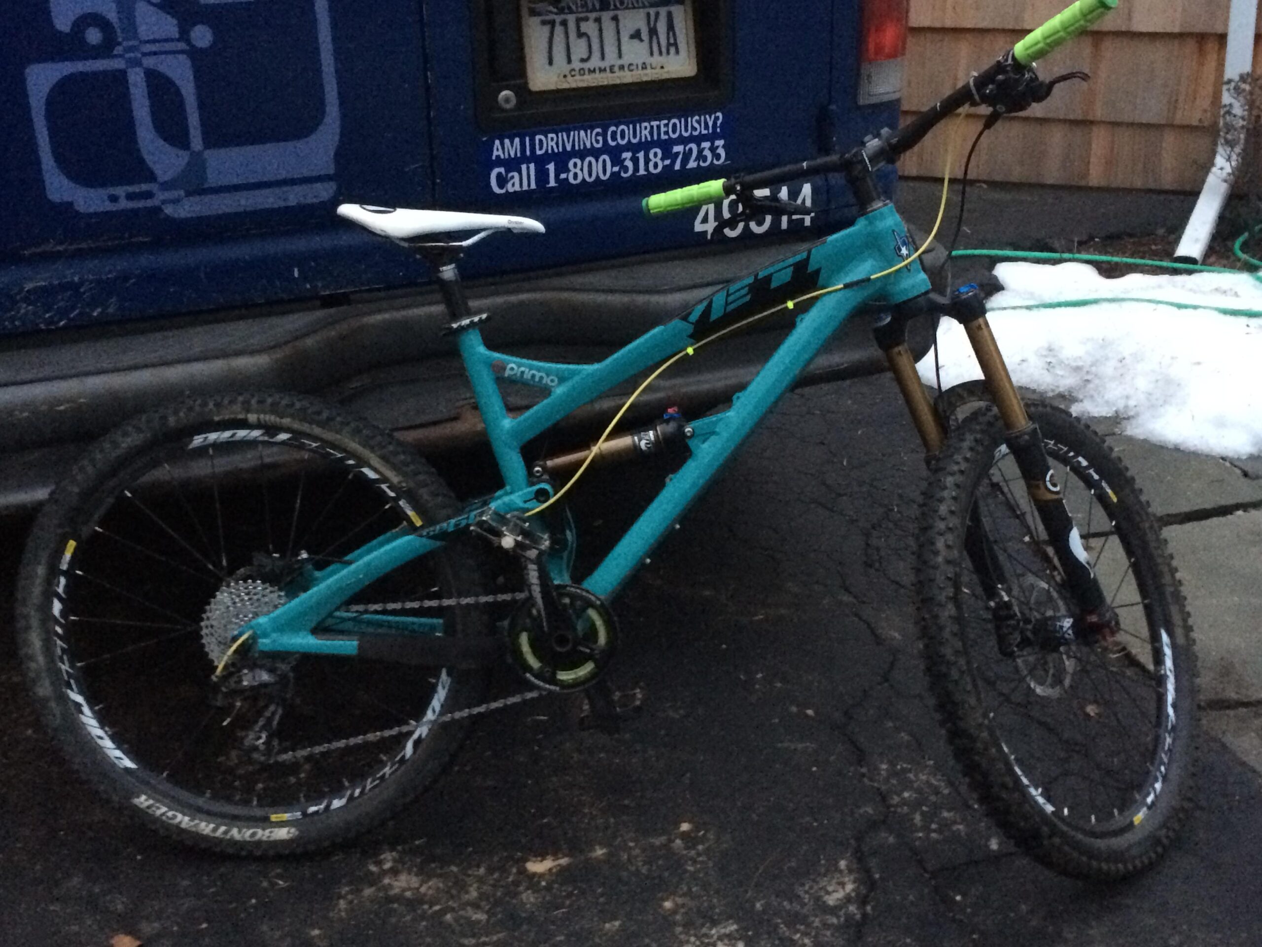Yeti SB66: A turquoise mountain bike with yellow cables, featuring a sturdy frame, front suspension, and thick tires, is parked on a cracked asphalt surface. Snow is visible in the background, indicating a cold environment.