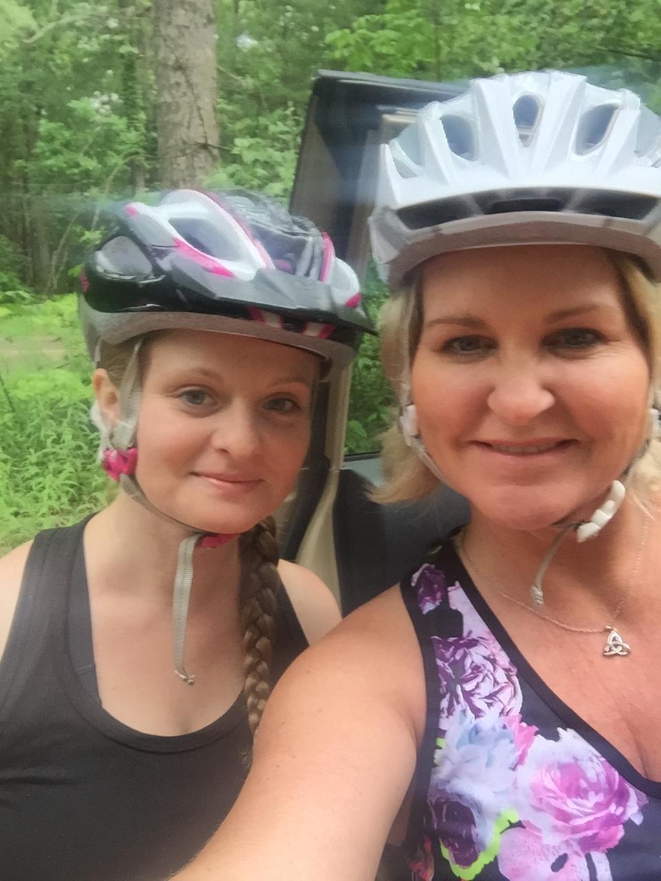 Specialized Rockhopper: Two women wearing bicycle helmets pose for a selfie in a lush green forest. The woman on the left has long, braided hair and wears a tank top, while the woman on the right has shoulder-length hair and is dressed in a sleeveless, floral-patterned top. They both smile, capturing a moment of joy in nature.