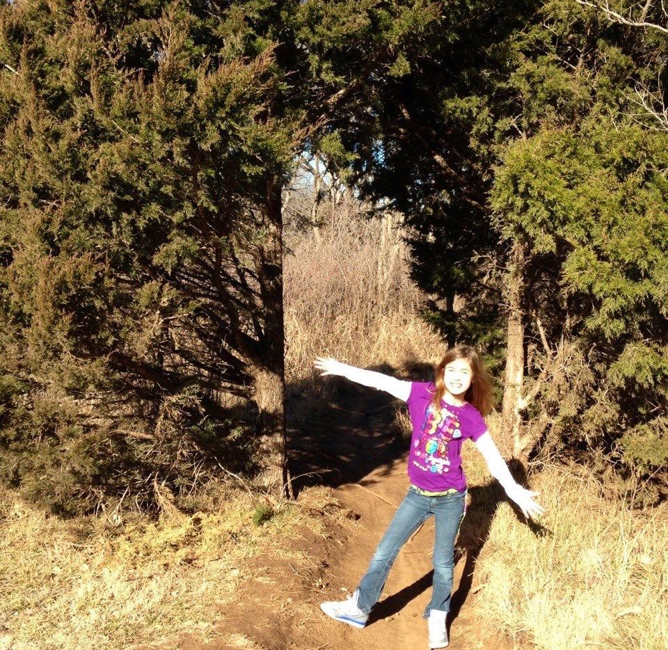 A young girl in a purple t-shirt and jeans stands happily on a dirt path surrounded by tall trees, with her arms outstretched as if enjoying the outdoors. Sunlight filters through the greenery, creating a cheerful and inviting atmosphere. Bluff Creek Trail mountain bike trail.