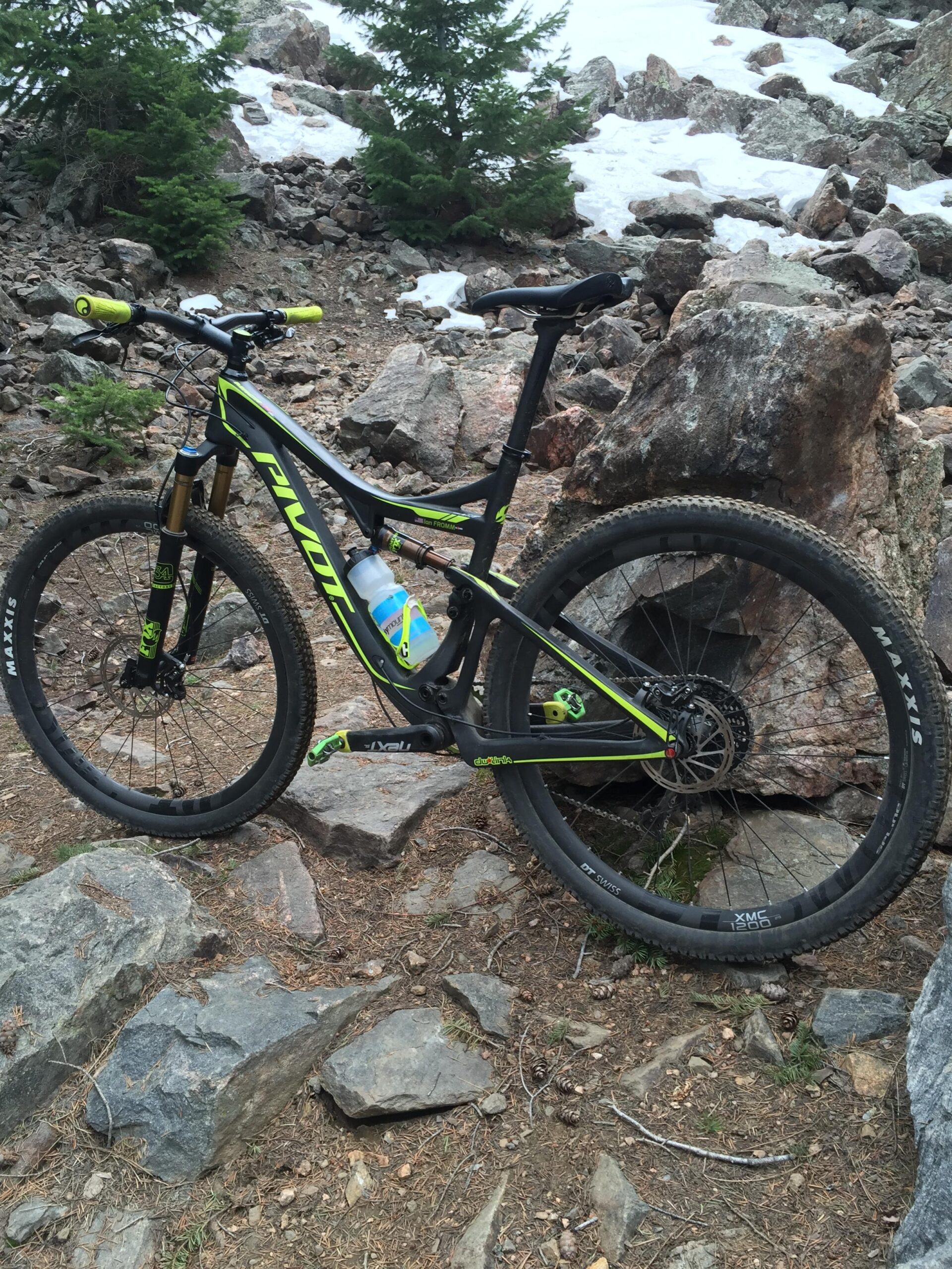 Pivot Mach 429SL Carbon: A mountain bike with a black and green frame is parked on a rocky trail surrounded by trees and patches of snow. The bike features yellow grips and a water bottle attached to the frame.