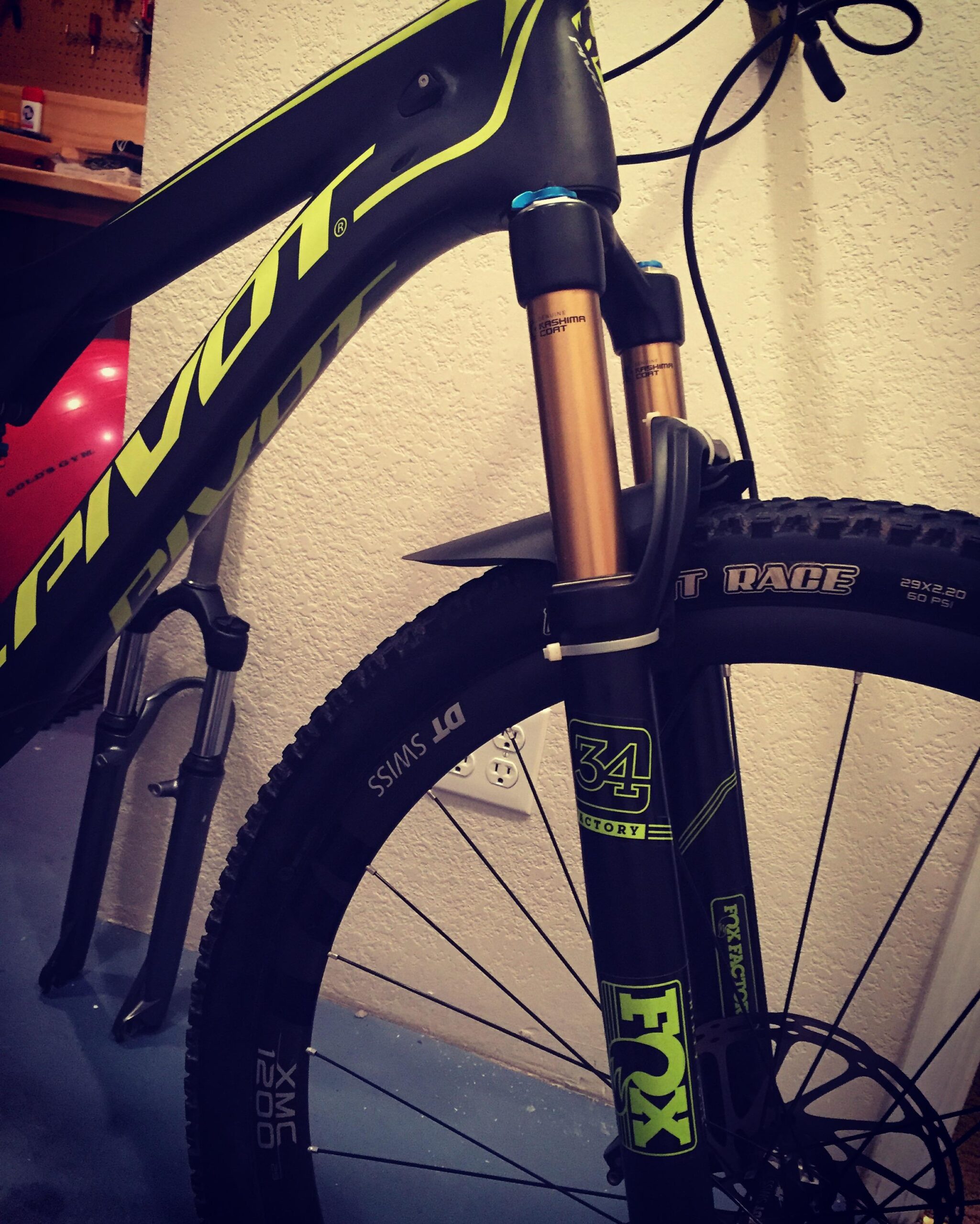 Pivot Mach 429SL Carbon: Close-up view of the front fork and wheel of a mountain bike, showcasing the black frame with bright green accents, highlighted markings on the fork, and the tire specifications. The background shows a textured wall and electrical outlet.