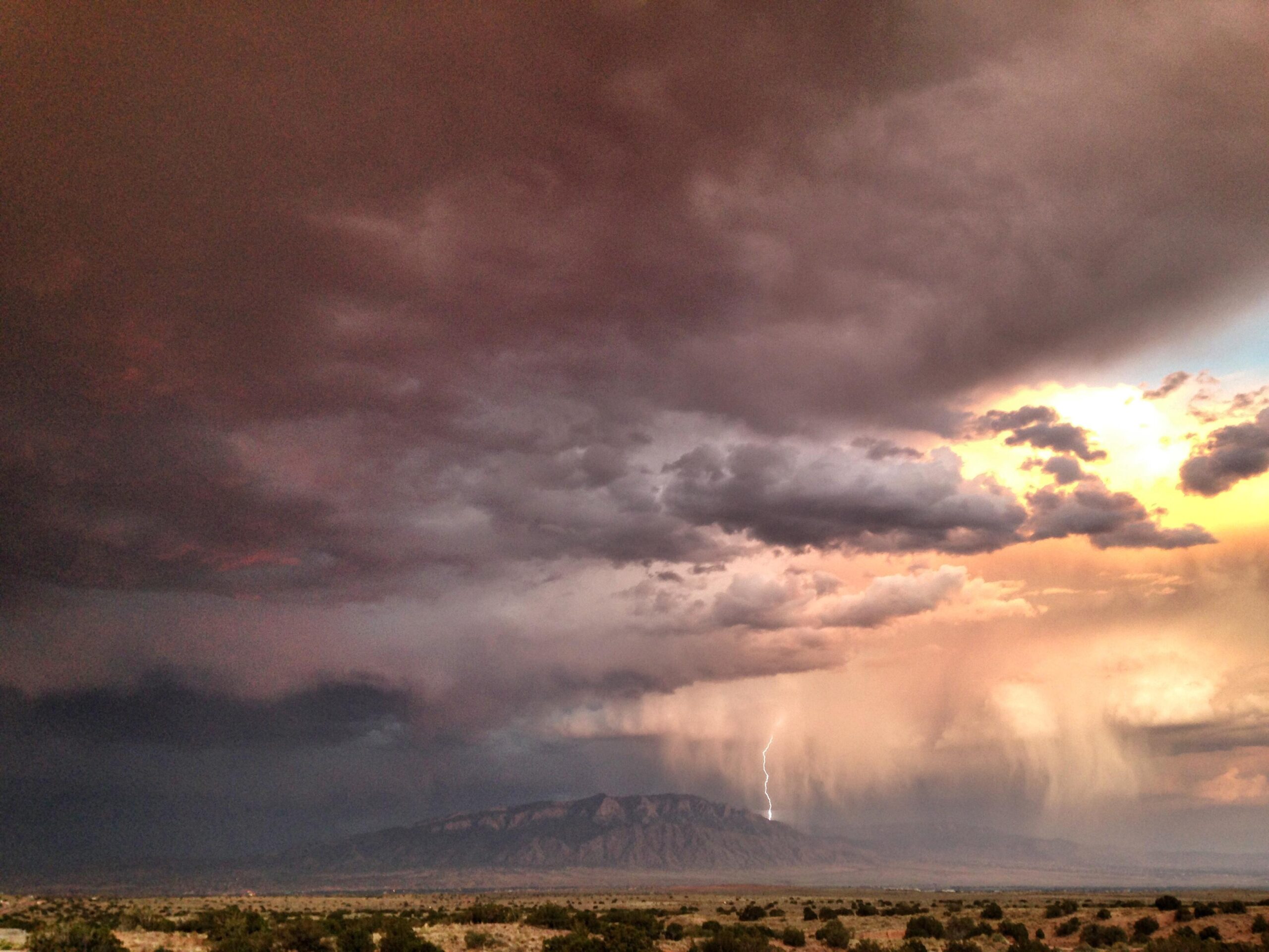 A dramatic landscape featuring dark storm clouds above a mountain range, with a flash of lightning striking in the distance. The sky displays a gradient of colors from deep gray to soft orange, indicating an approaching storm, while rain can be seen falling from the clouds. The foreground includes sparse vegetation typical of a desert setting. Mariposa Fat Bike Trails mountain bike trail.