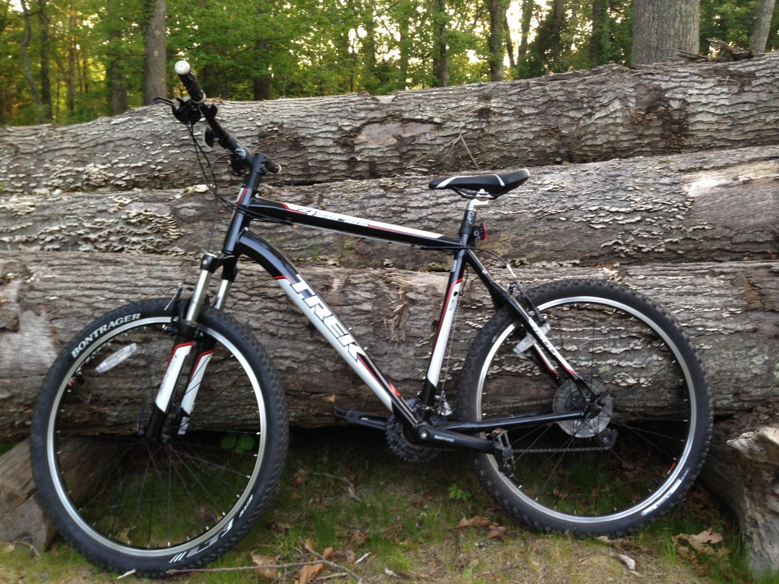 Trek 4300: A black and silver mountain bike is positioned next to a large fallen tree trunk in a wooded area. The bike features distinct Bontrager tires and a sleek frame with a prominent Trek logo. Surrounding greenery and the texture of the log enhance the natural setting.