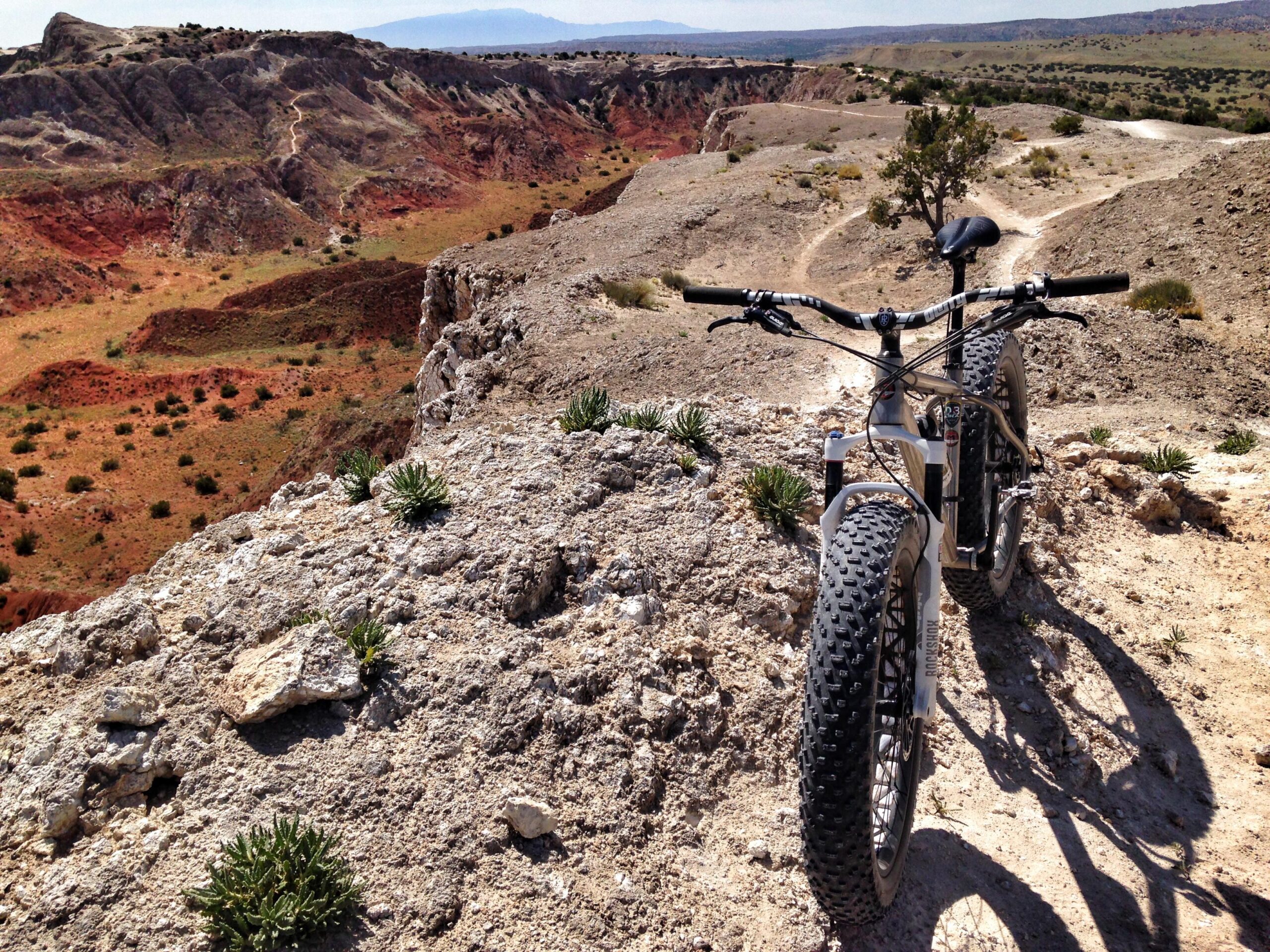 A fat tire bike resting on a rocky ledge overlooking a colorful desert landscape. The terrain features red and orange earth with sparse vegetation, and a winding trail can be seen in the distance against a backdrop of rolling hills and mountains under a clear blue sky. White Ridge Bike Trails mountain bike trail.