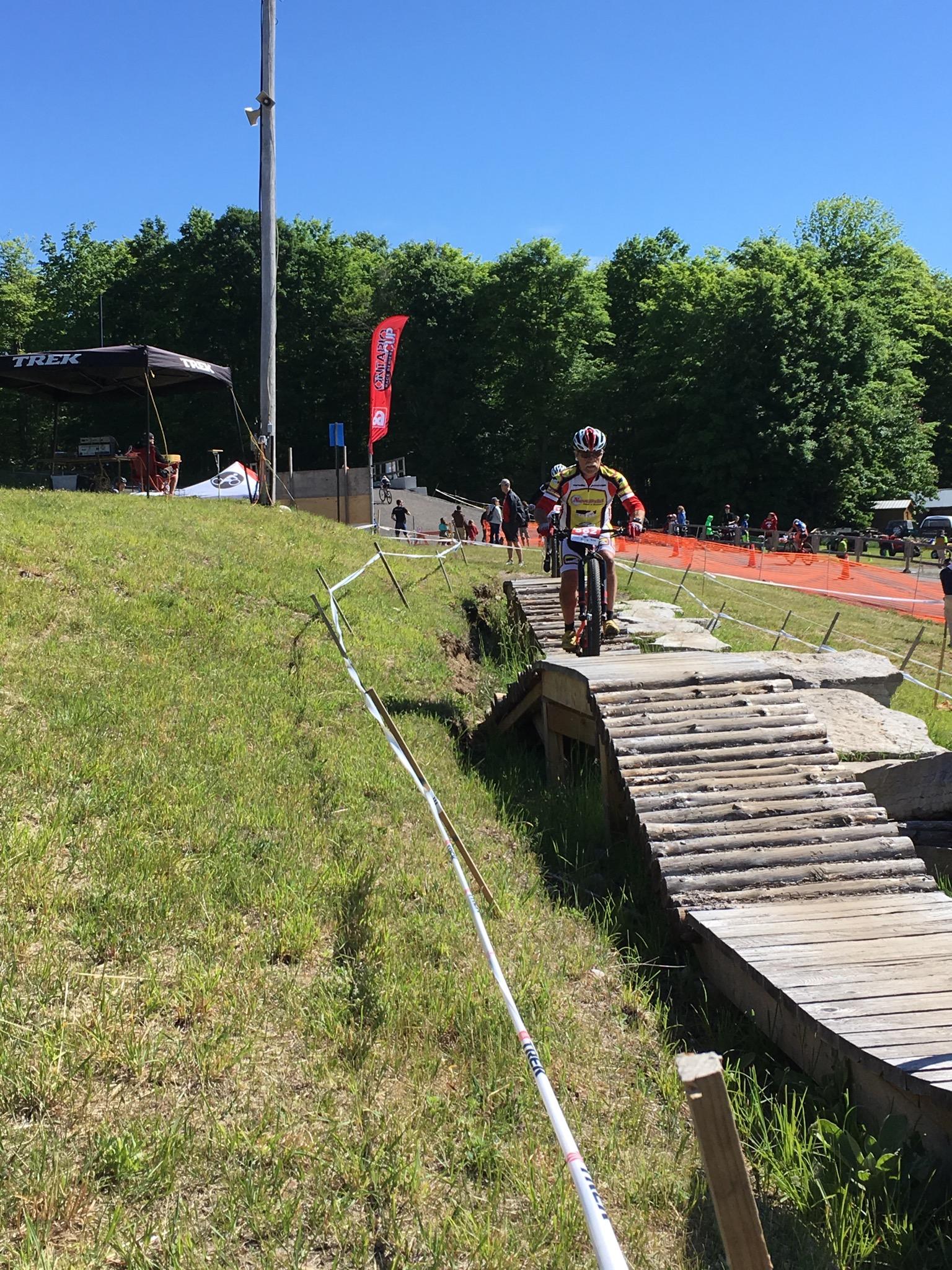 A mountain biker riding on a wooden bridge in a green grassy area, surrounded by trees and a clear blue sky. In the background, spectators are gathered along the course, and banners are visible, indicating a cycling event. A tent with the logo "TREK" can be seen on the left side of the image. Hardwood Ski and Bike mountain bike trail.