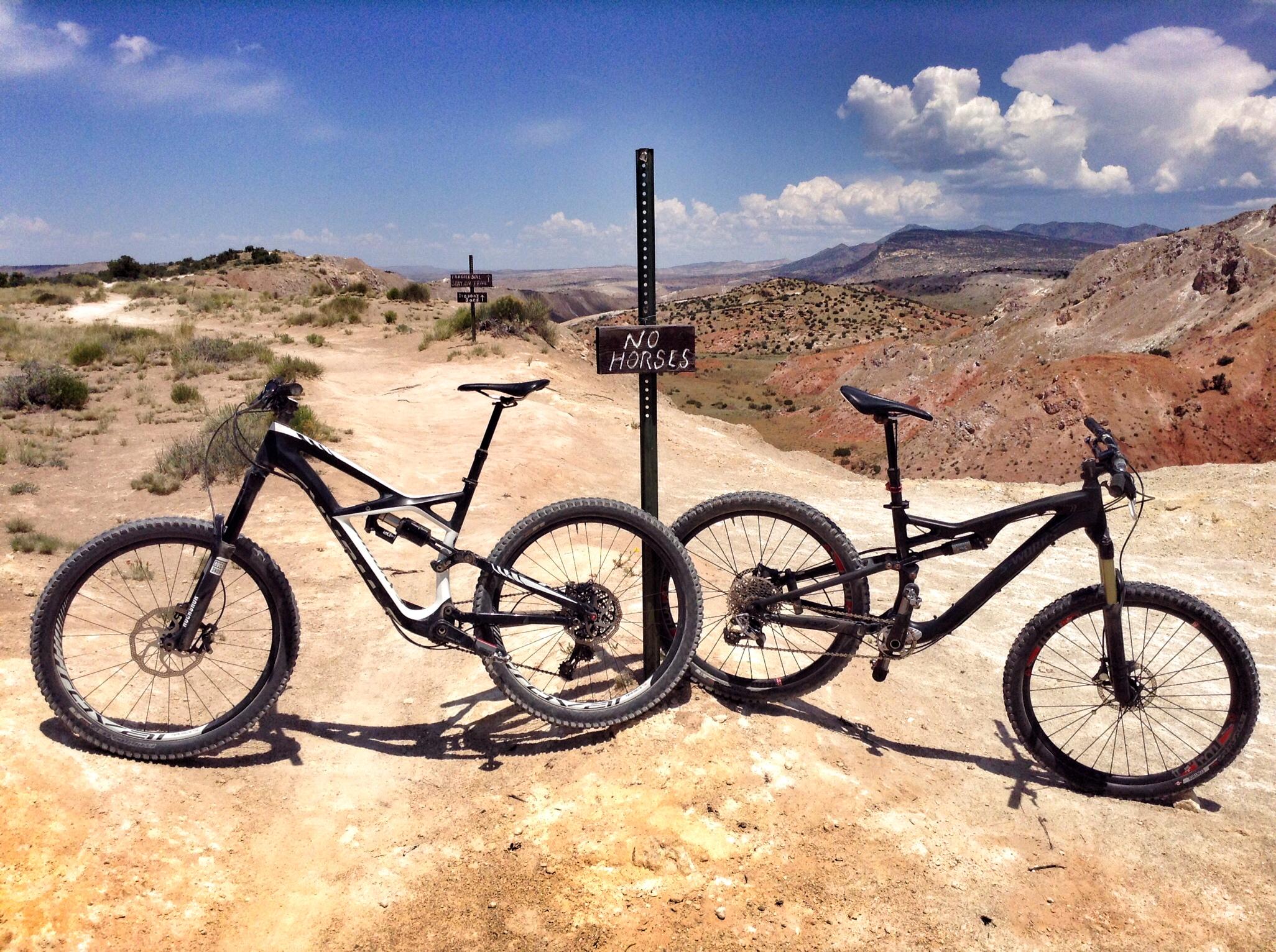Two mountain bikes positioned near a "No Horses" sign on a dirt trail, surrounded by rocky terrain and a clear blue sky with scattered clouds in the background. The landscape features rolling hills and vibrant natural colors, typical of an outdoor biking environment. White Ridge Bike Trails mountain bike trail.