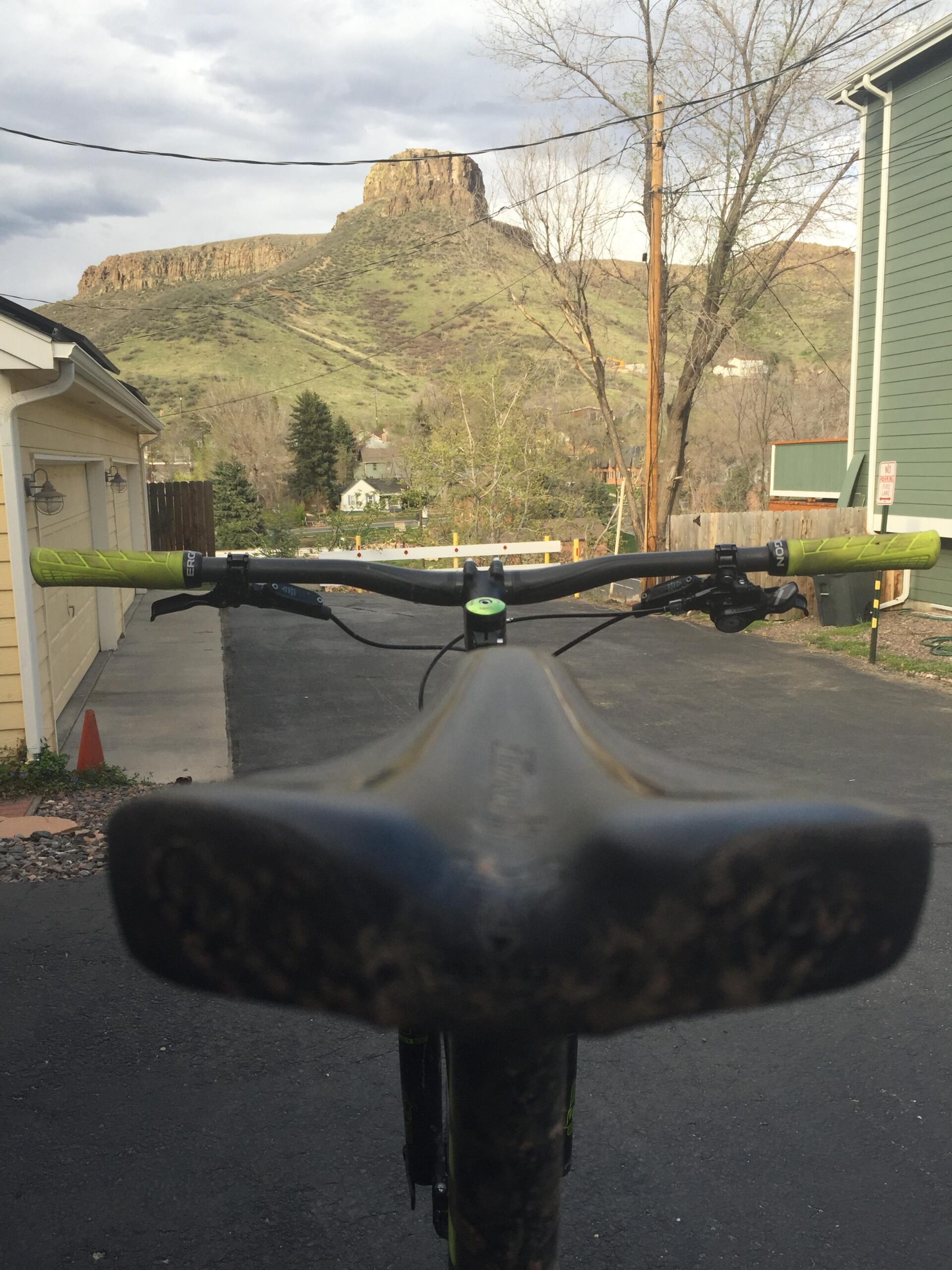 Pivot Mach 429SL Carbon: A view from the seat of a mountain bike, looking down the handlebars towards a prominent rock formation and hillside in the background. The setting features a paved driveway and residential buildings on either side, with trees and power lines adding context to the rural landscape under a cloudy sky.
