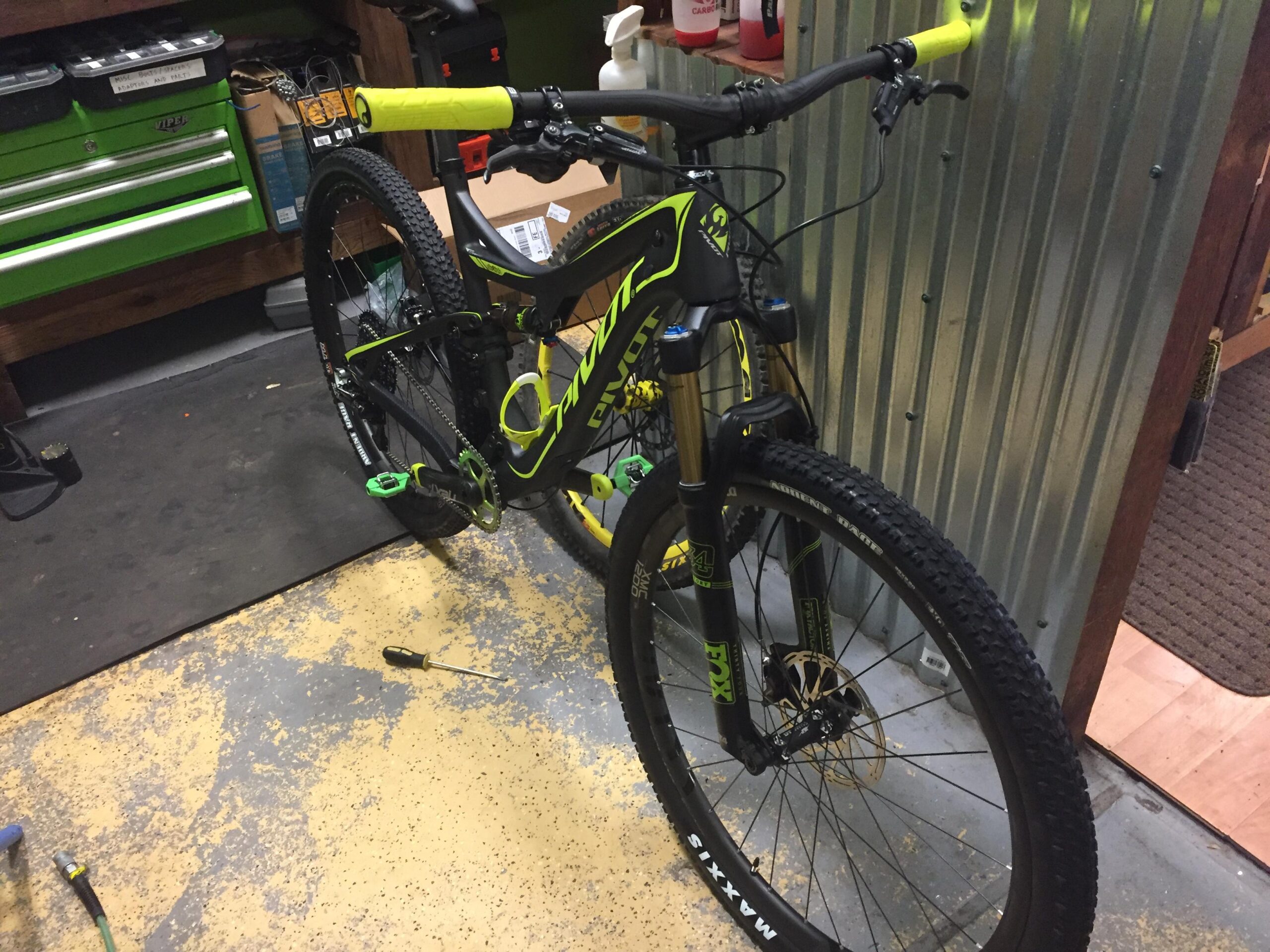 Pivot Mach 429SL Carbon: A brightly colored mountain bike with a black and neon green design is positioned in a garage setting. The bike features thick tires and a front suspension, resting on a textured floor with a few tools and equipment visible in the background, including a green tool chest and various items on shelves.