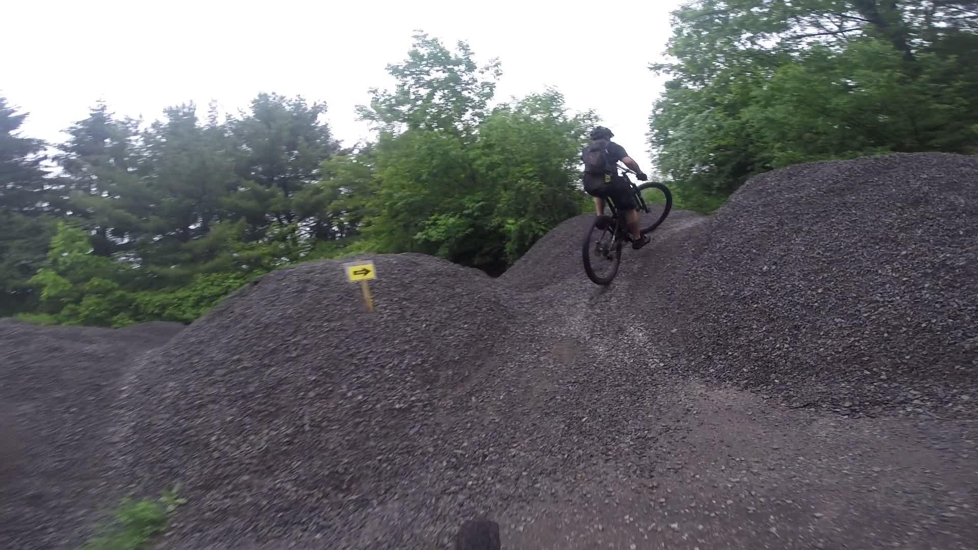 A mountain biker skillfully navigates a gravel pile at an outdoor biking park, catching air as they ride over a mound surrounded by lush green trees. A directional sign indicates the path ahead. Stewart State Forest mountain bike trail.