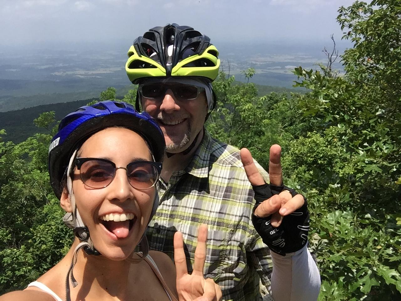 Trek Fuel EX 8: A smiling man and woman wearing helmets pose for a selfie outdoors, with a scenic view of lush green mountains in the background. Both are making peace signs with their fingers, and the woman is sticking out her tongue playfully. The sun is shining, creating a cheerful atmosphere.