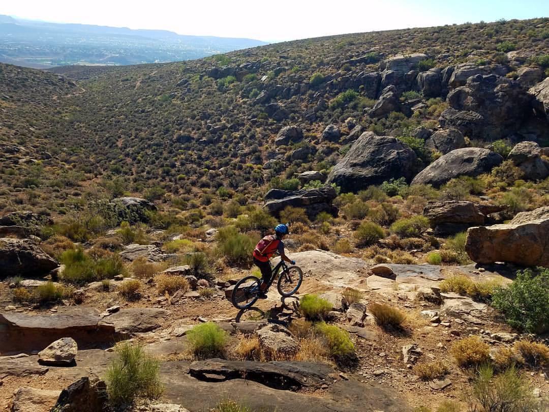A person riding a mountain bike on a rocky trail surrounded by sparse vegetation and hills, with a distant landscape visible in the background under a clear sky. Barrel Roll mountain bike trail.