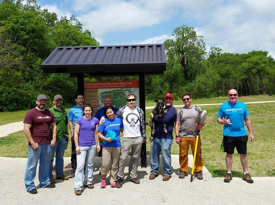 A group of ten people, including a man holding a dog, stand in front of a sign for the Bluff Creek Park Mountain Bike Trail. The individuals are smiling and dressed in casual outdoor clothing, surrounded by greenery and a clear blue sky. Some are holding tools, indicating a community event or maintenance activity. Bluff Creek Trail mountain bike trail.