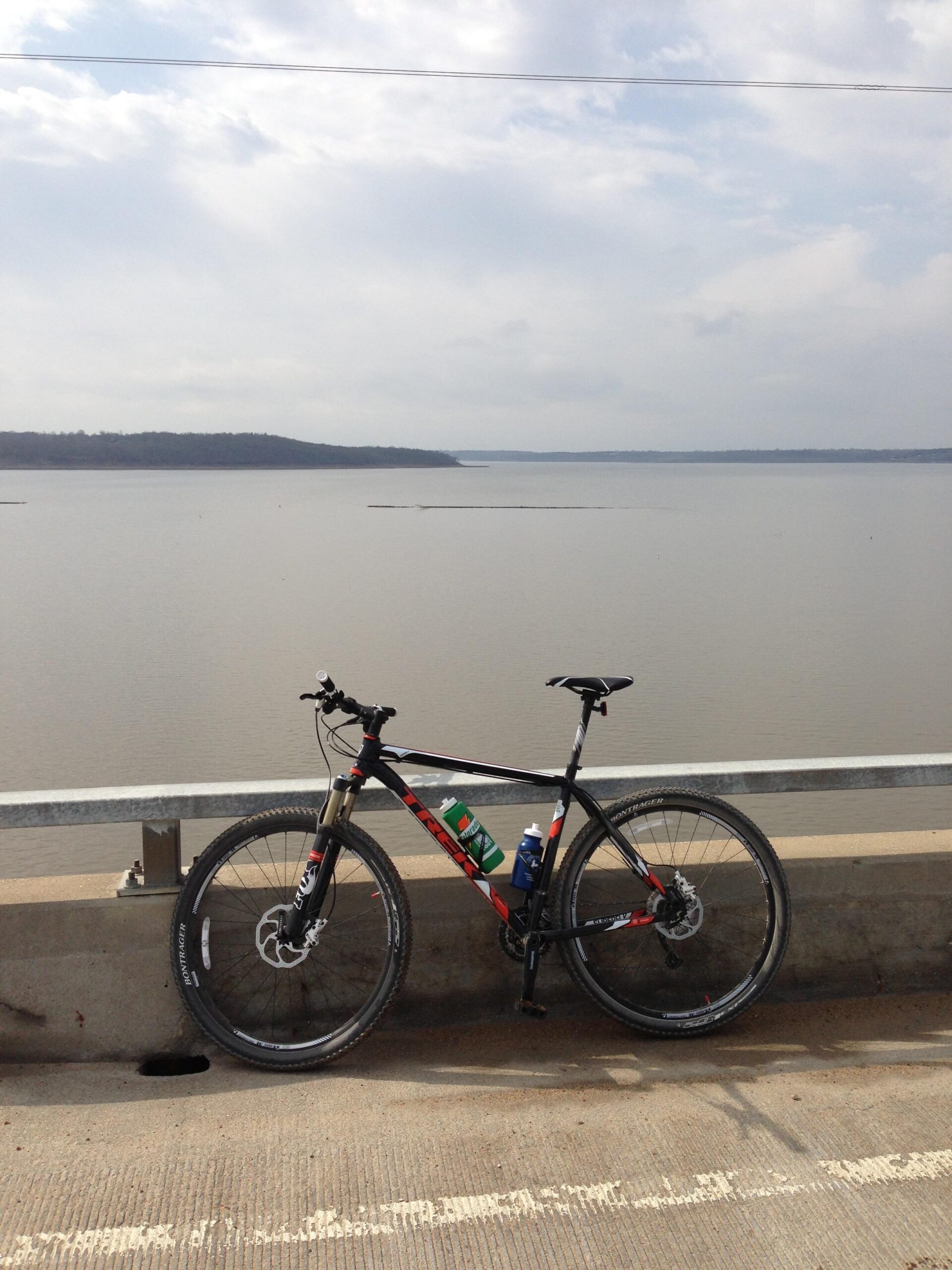 Trek Superfly AL: A black mountain bike with red and white accents is parked on a concrete barrier overlooking a calm body of water. The bike has a water bottle holder with a blue bottle and a green container. The background features a cloudy sky and a distant forested shoreline.