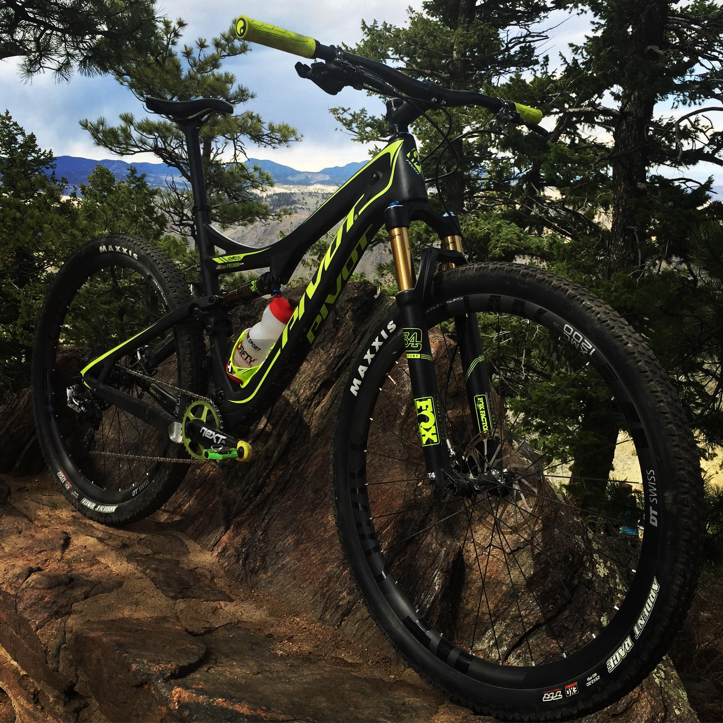 Pivot Mach 429SL Carbon: A mountain bike with bright green and black accents is positioned on a rocky surface amid trees, with a backdrop of mountains under a cloudy sky. A water bottle is mounted in the bike's frame.